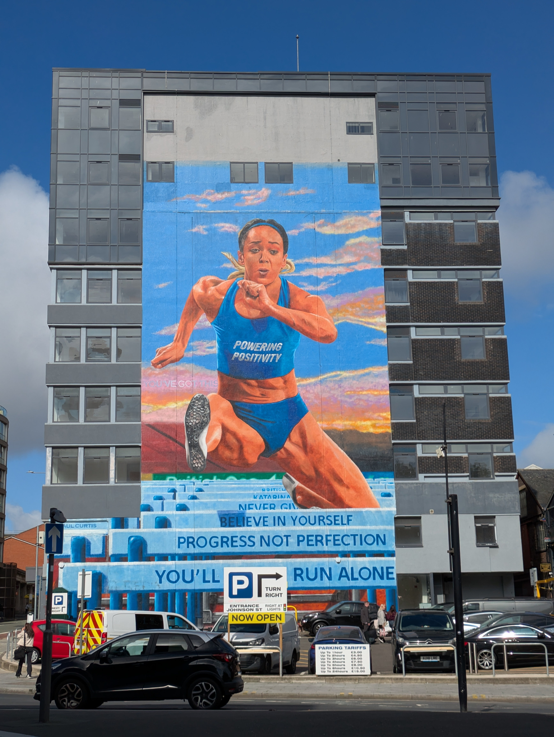 A large mural of KJT in mid-jump wearing a blue outfit with the words POWERING POSITIVITY is painted on the side of a building, accompanied by motivational phrases about belief, progress, and not running alone.