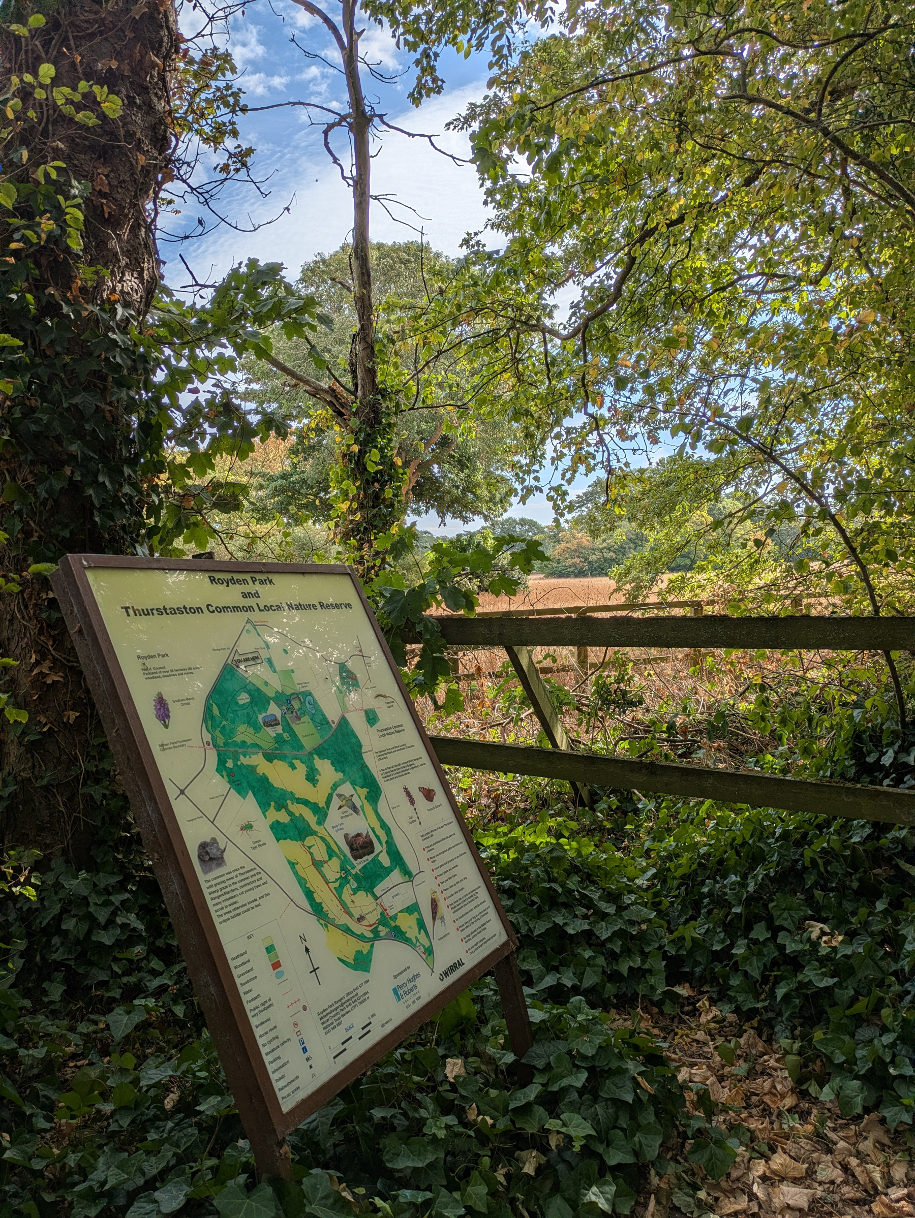 A wooden signboard displaying a map of Royden Park and Thurstaston Common Local Nature Reserve stands amidst green foliage and trees near a rustic wooden fence under a partly cloudy sky.