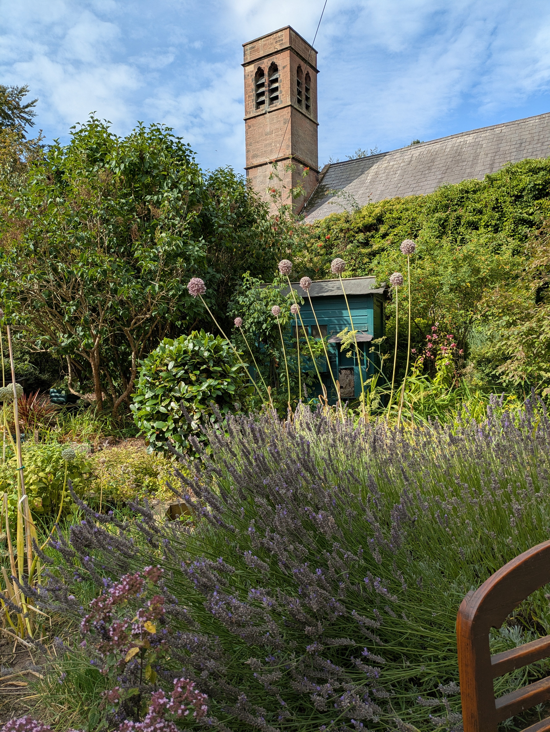 A tall brick tower rises behind a lush garden filled with various plants, flowers, and greenery under a partly cloudy sky.