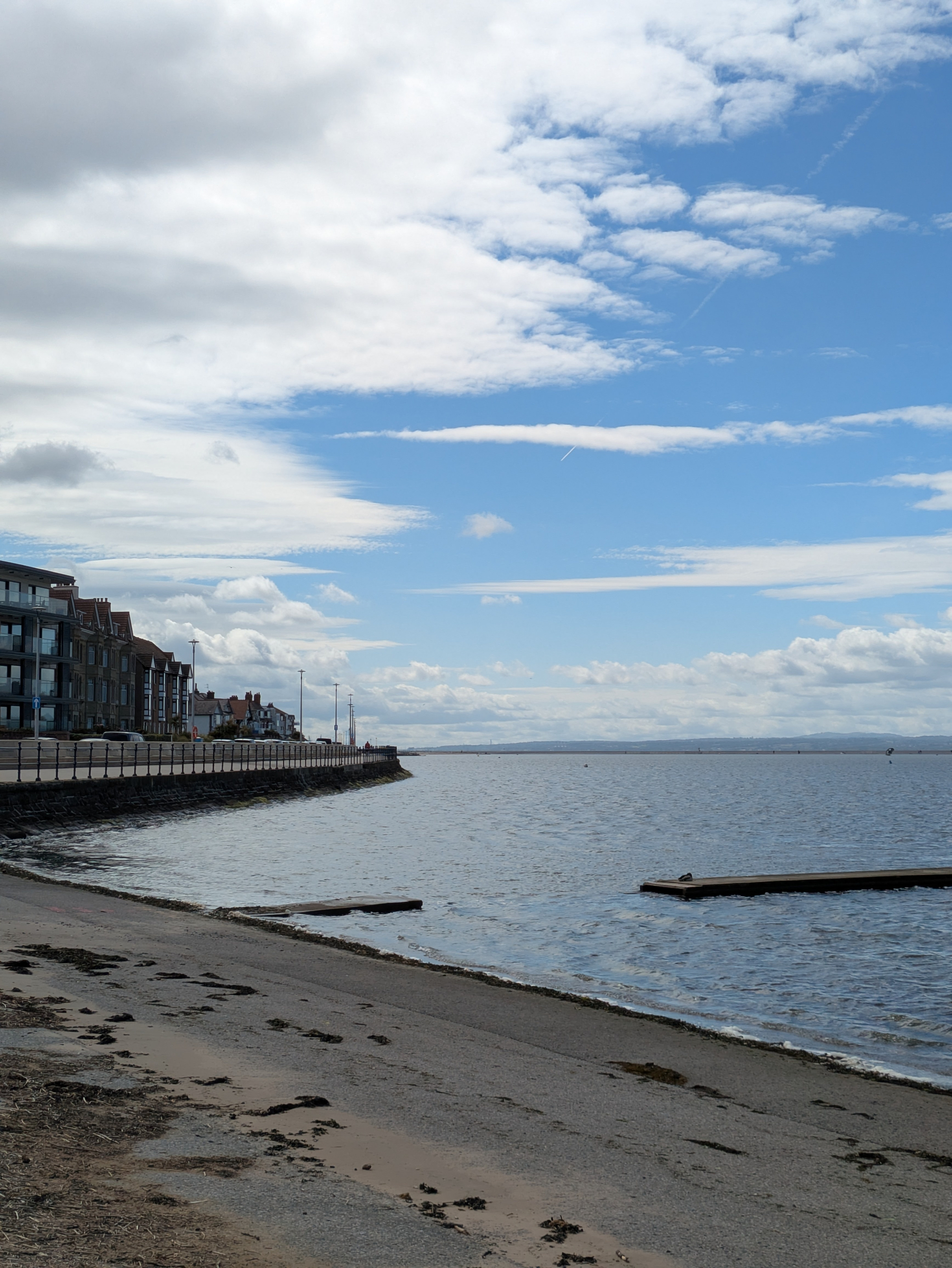 A serene coastal scene features calm waters, a sandy beach, and a row of buildings along the shoreline under a partially cloudy sky.