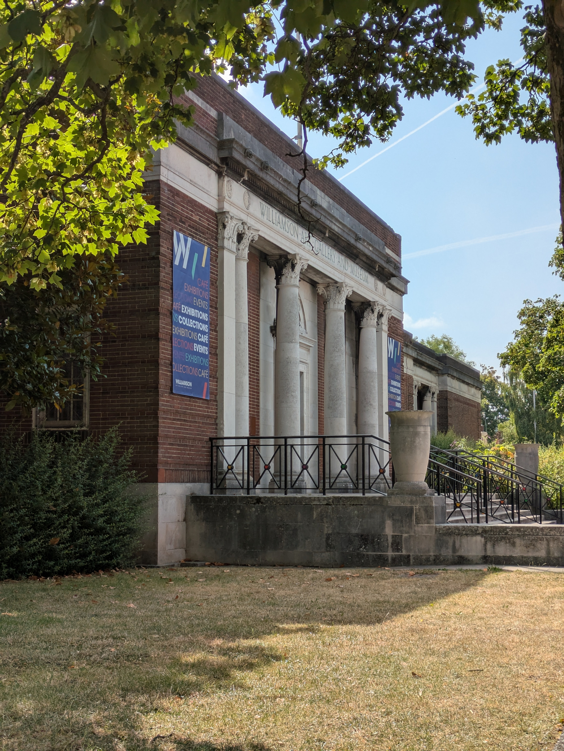 A historic brick building with classical white columns and a banner is partially framed by leafy green trees.