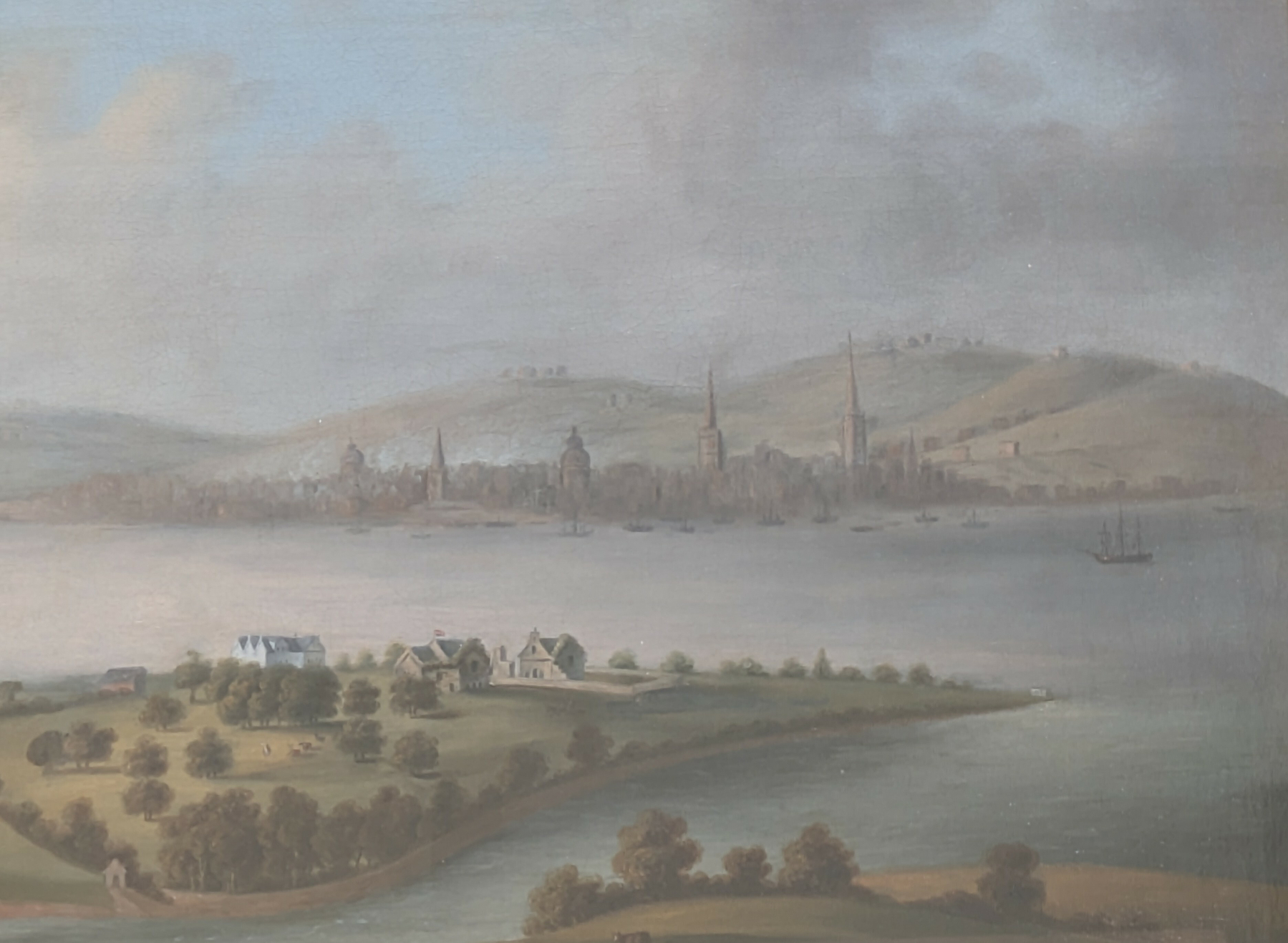A pastoral landscape features a river with a small settlement in the foreground and a cityscape with tall spires and hills in the background.