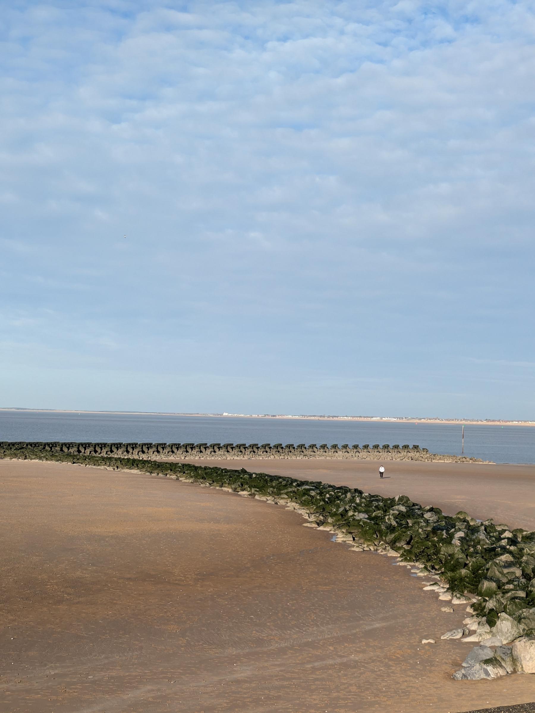 A sandy beach with a curved rock barrier extends into the calm sea under a partly cloudy sky.