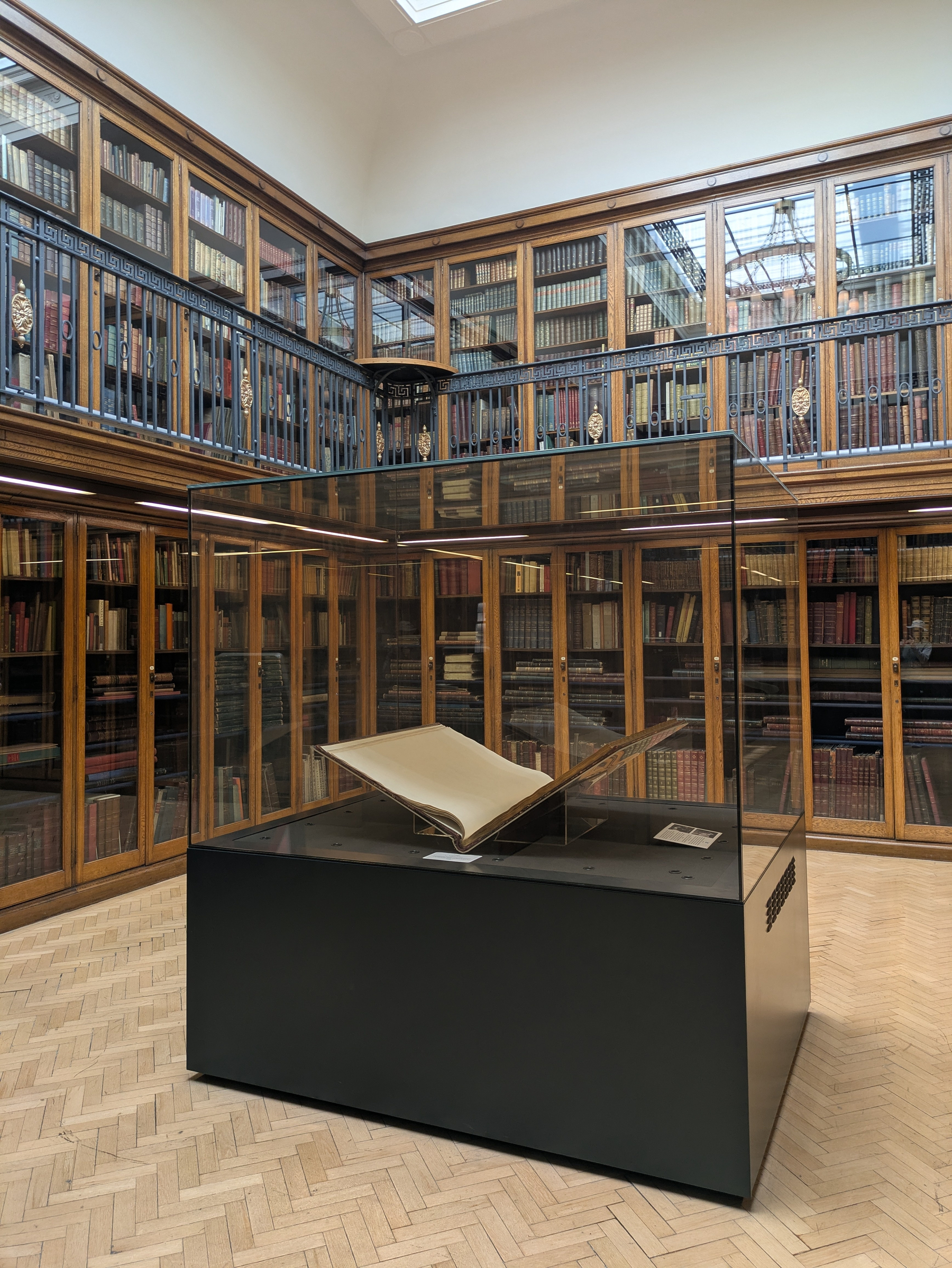 A large, open book is displayed in a glass case in the center of a library with tall wooden bookshelves filled with books.