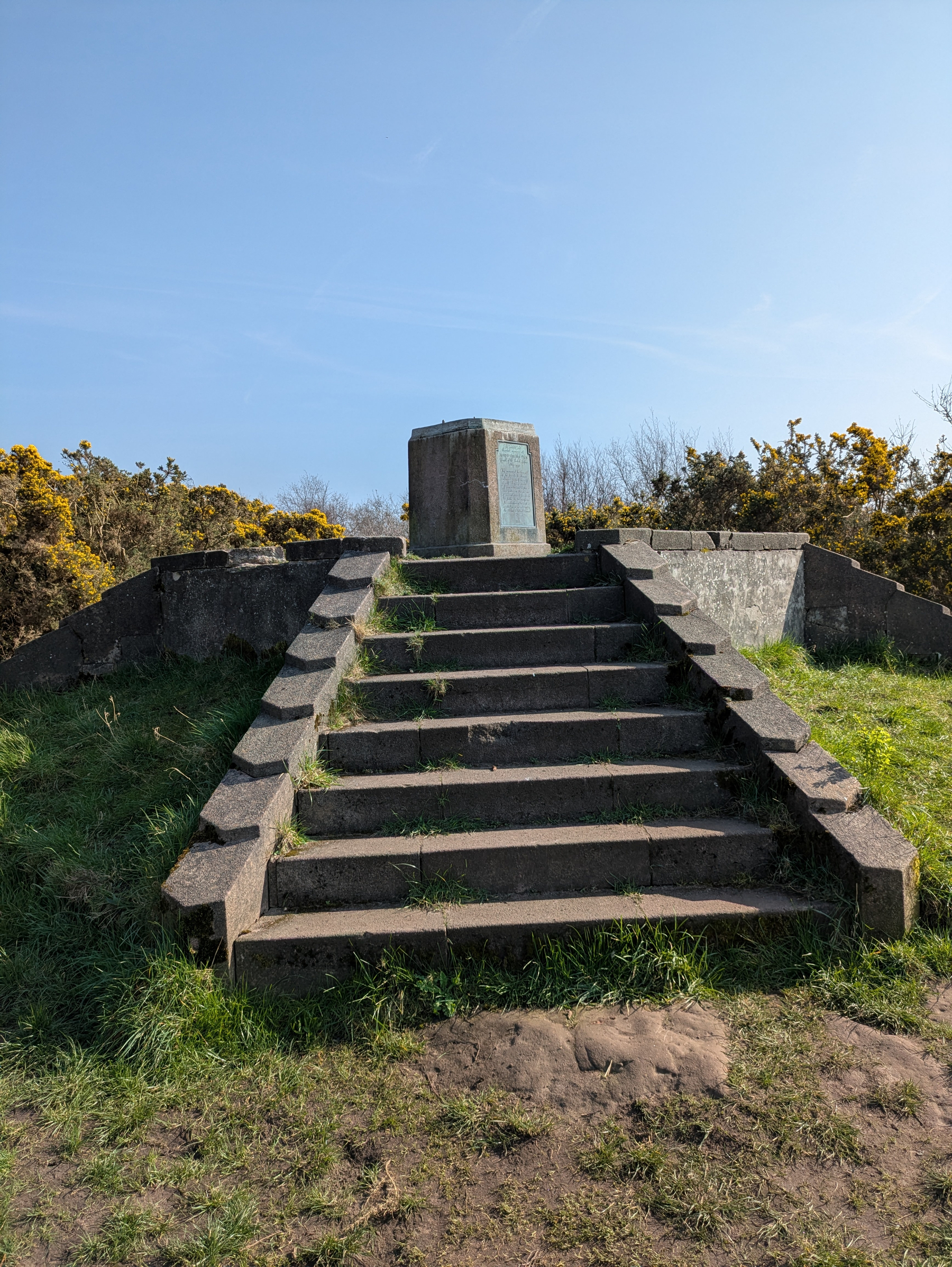A stone monument sits at the top of a set of stone steps, surrounded by grass and bushes under a clear blue sky.