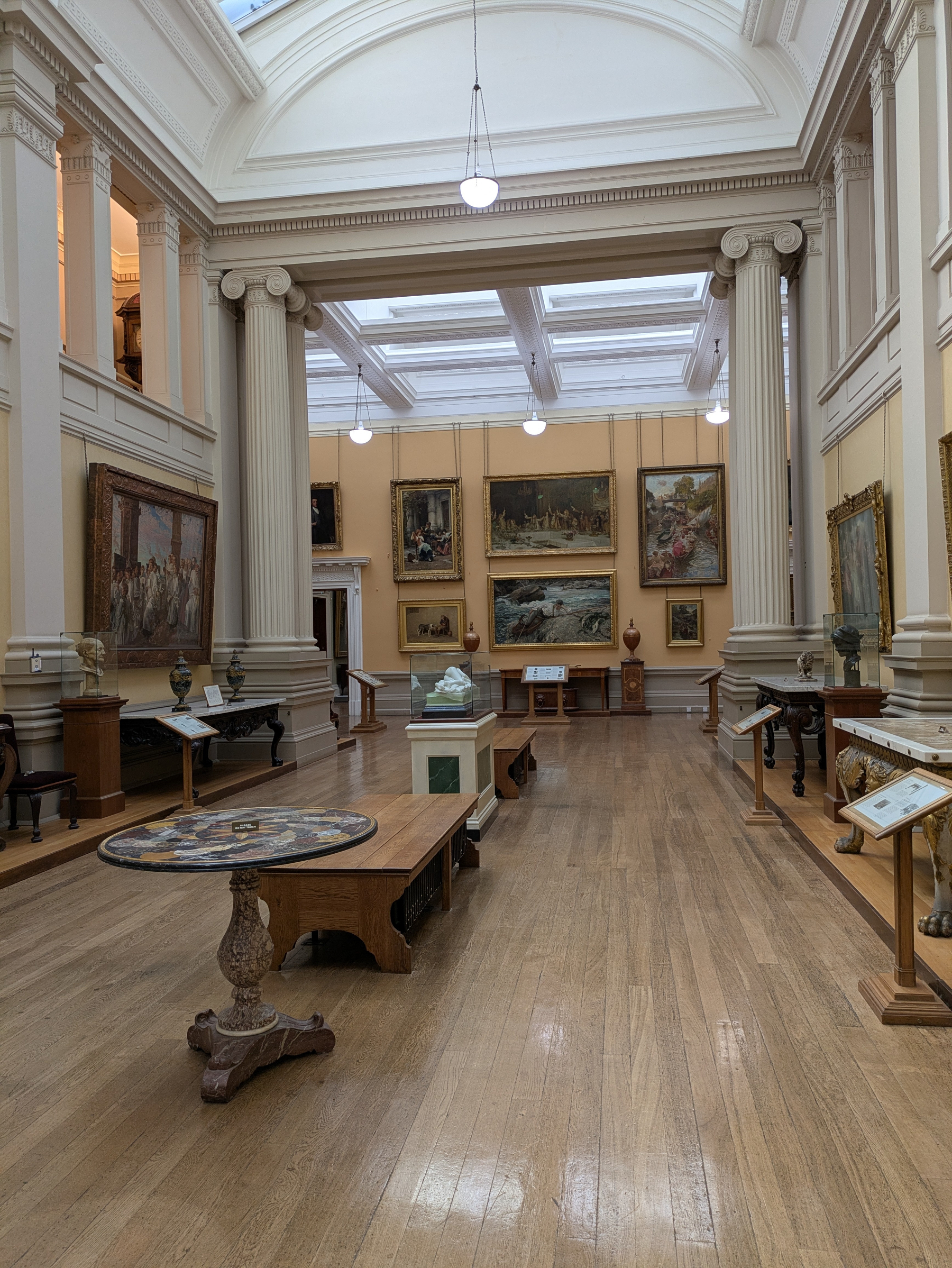 A gallery hallway is adorned with classical paintings, sculptures, and elegant furniture beneath a high vaulted ceiling with skylights.