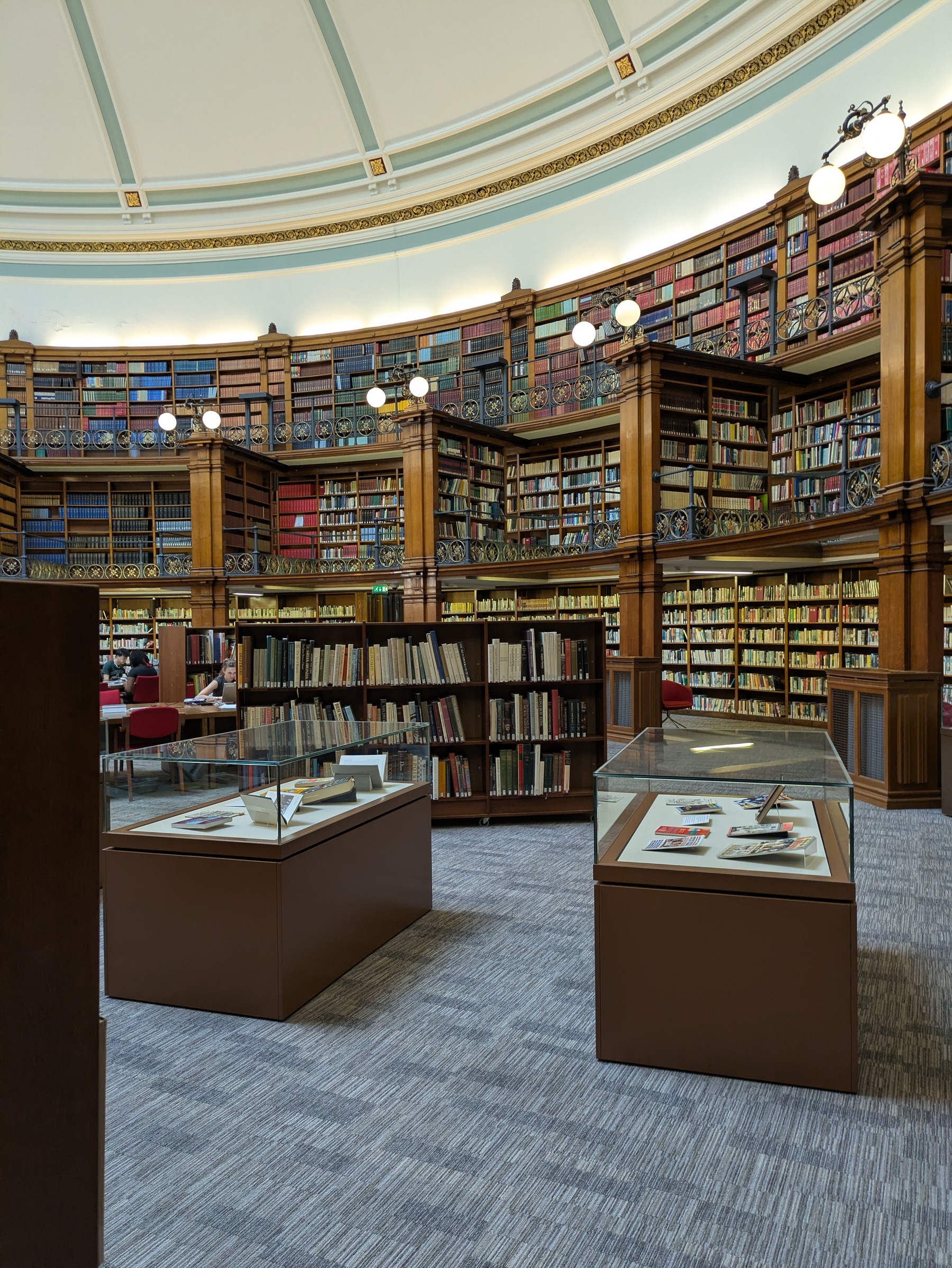 A large, circular library room with wooden bookshelves filled with books, display cases, and a domed ceiling is shown.