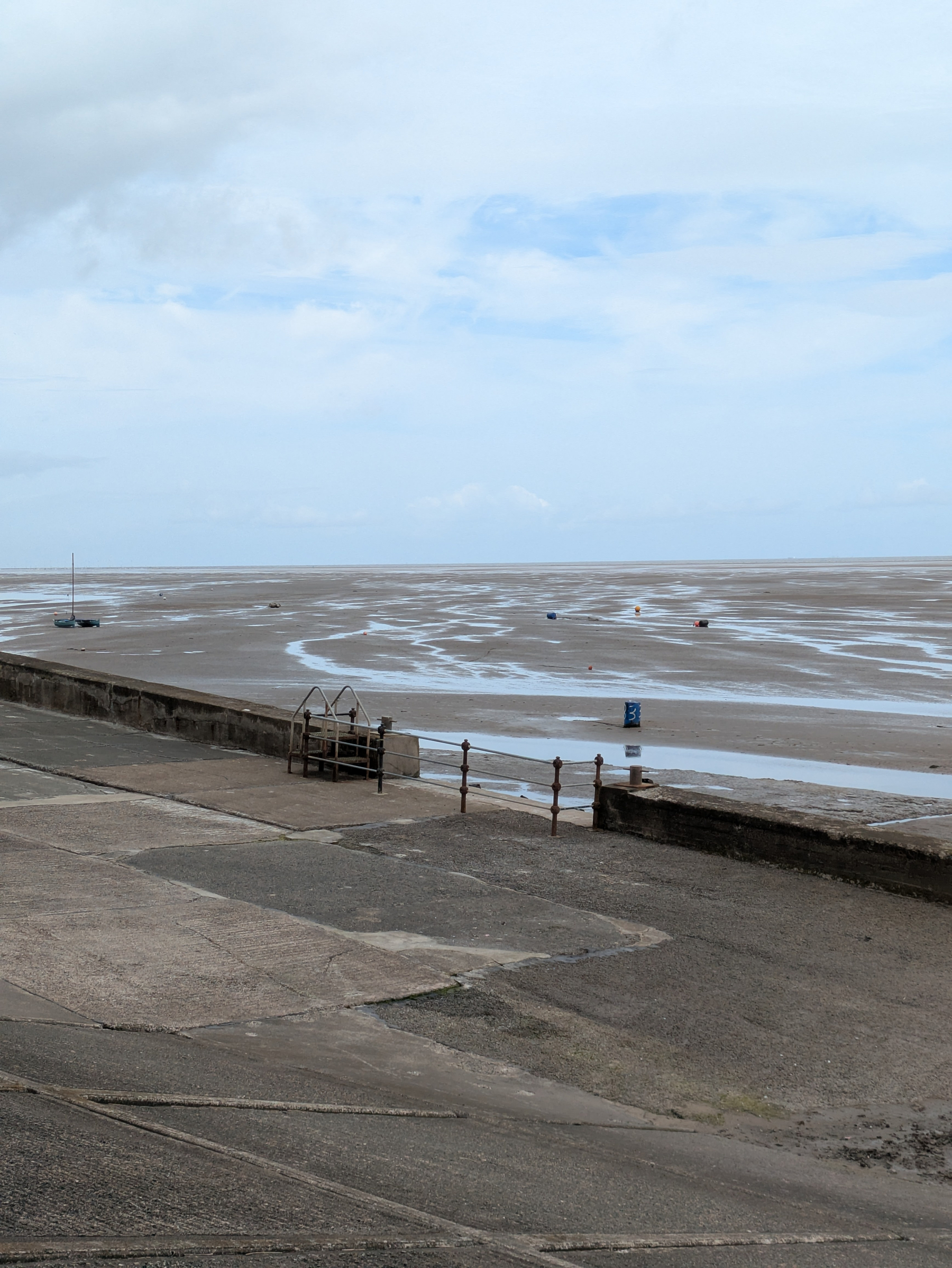 A sandy beach with scattered small pools of water stretches out to meet the cloudy sky, with a few boats visible in the distance and a paved area in the foreground.