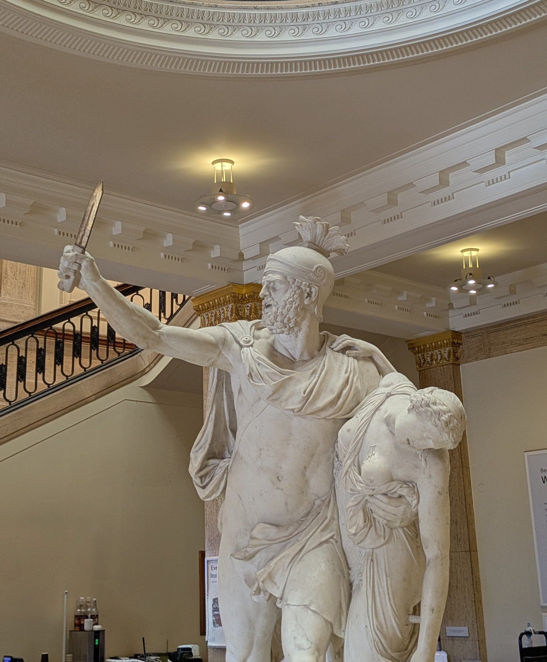 A classical statue depicts a muscular figure holding a dagger while supporting another figure in a grand interior setting.