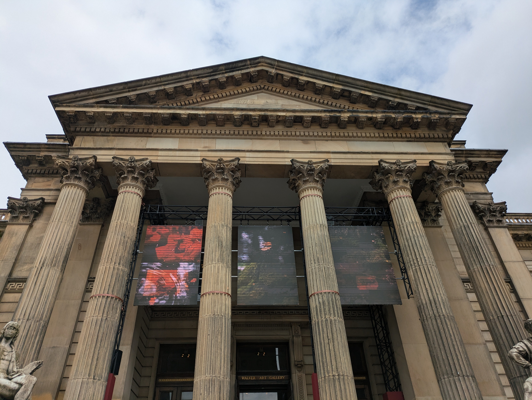 A neoclassical building features tall columns and two large digital screens displaying colorful images under a cloudy sky.
