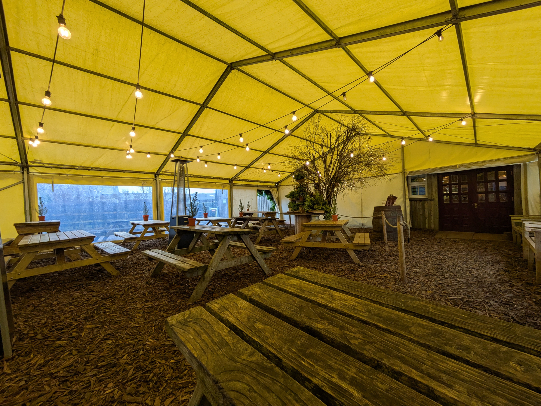 Under the canopy at Benty's with wooden picnic benches, wood chippings on the floor, propane heaters, strings of lights and a central rustic feature. Doors to the servery on right. 