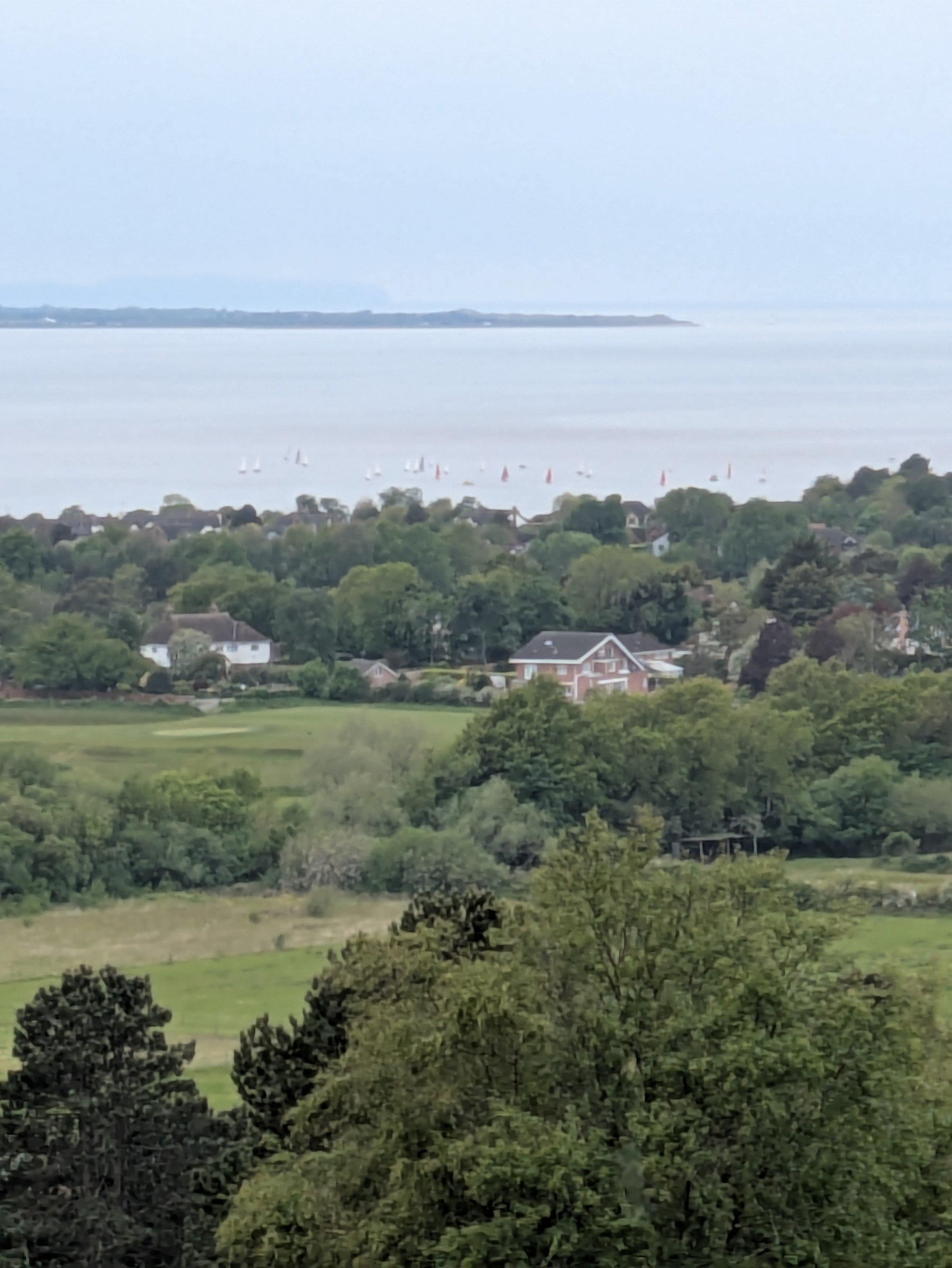 A serene coastal landscape features a distant view of the sea with sailboats, surrounded by lush greenery and houses.
