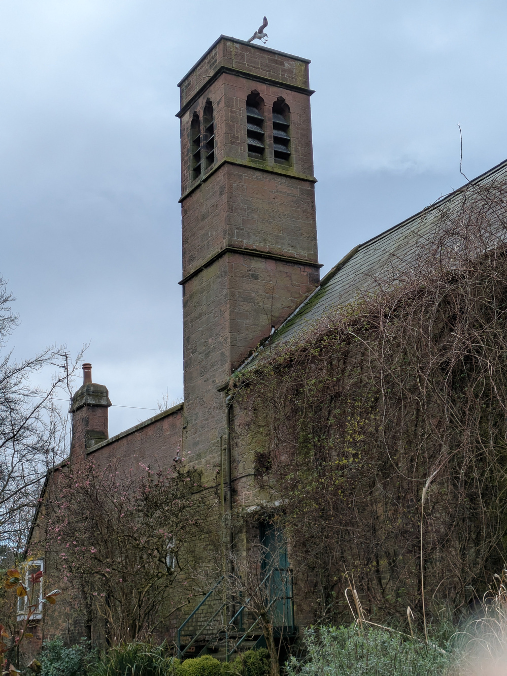 A stone building with a tall, narrow bell tower is surrounded by bare trees and overcast skies.