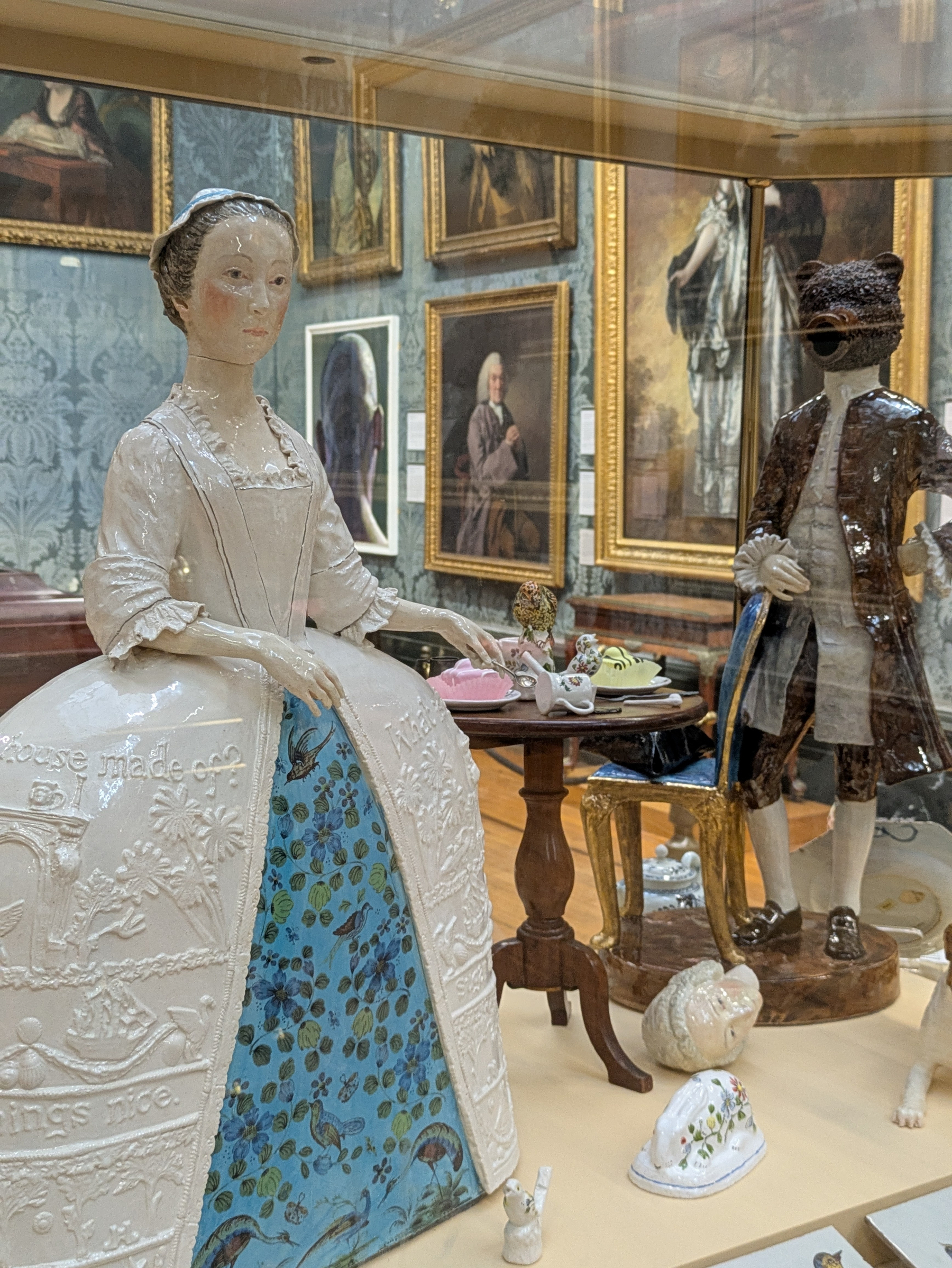 A museum exhibit displays ceramic figures, including a detailed statue of a woman in a dress with floral patterns and a figure of a person with an animal head, surrounded by other ceramic items and framed paintings on the walls.