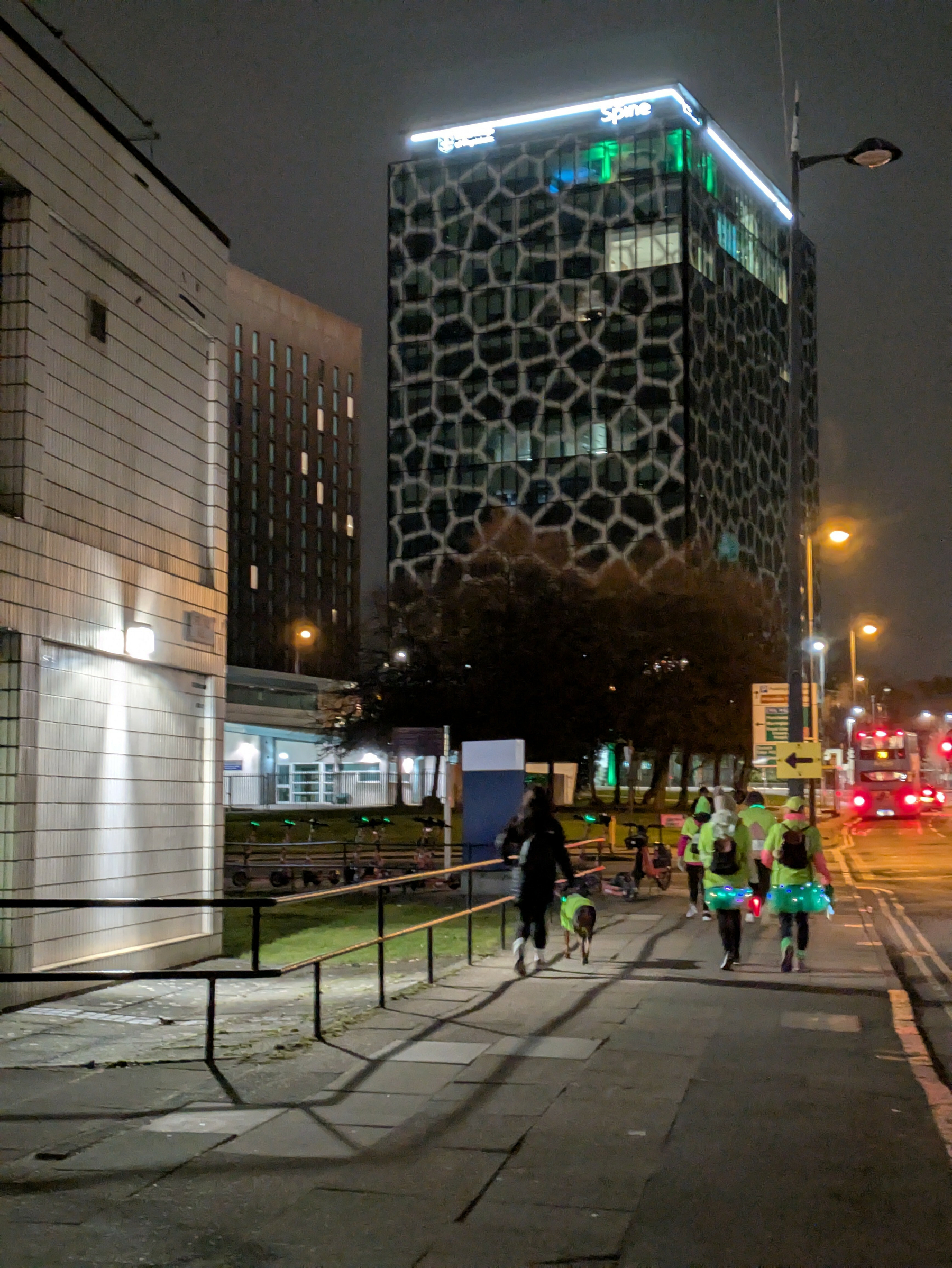 A group of people dressed in neon clothing and tutus jogs down a city sidewalk at night near a tall, uniquely patterned building.