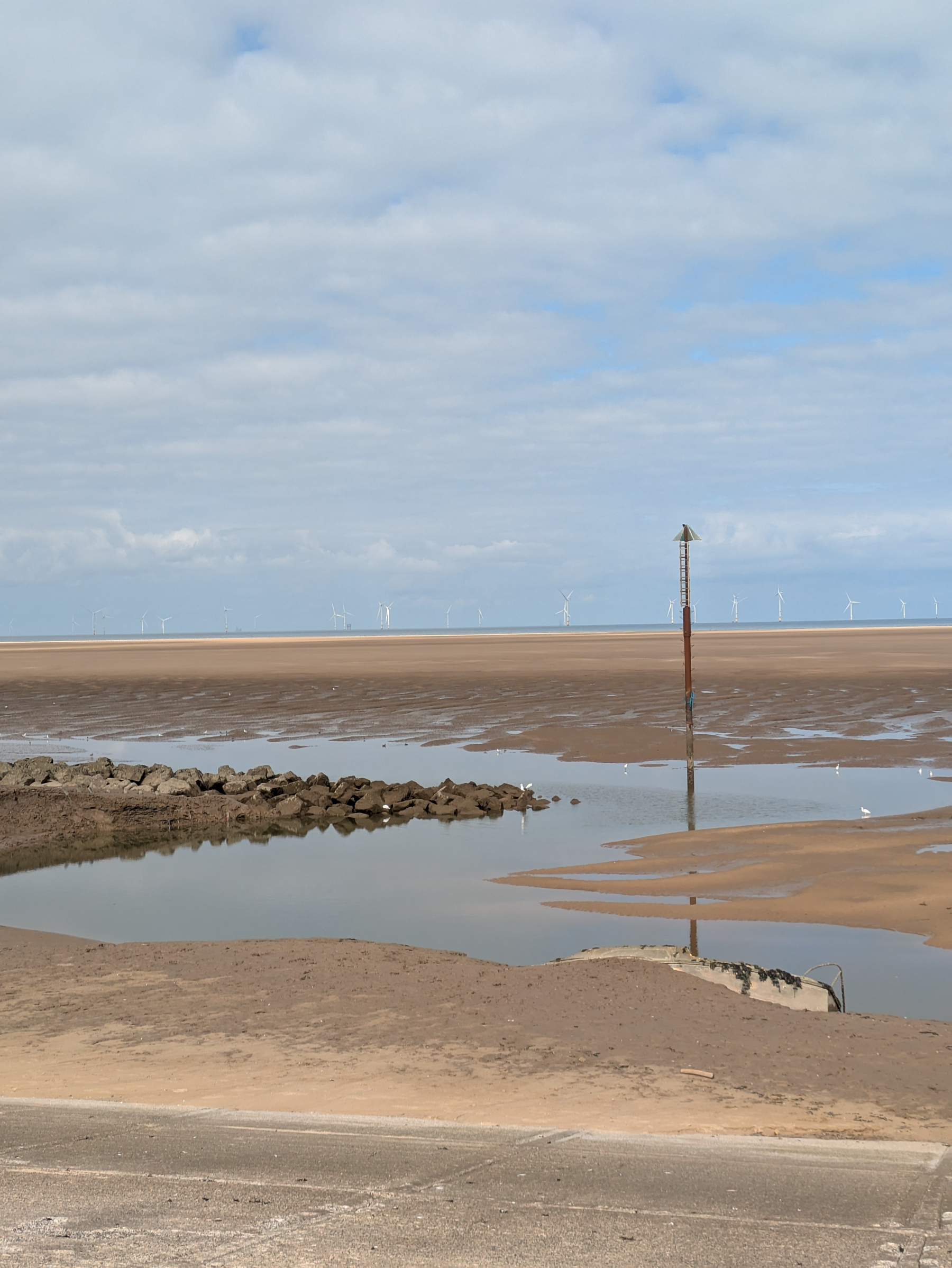 A calm beach landscape with shallow water pools, a marker post, and wind turbines visible on the horizon under a partly cloudy sky.