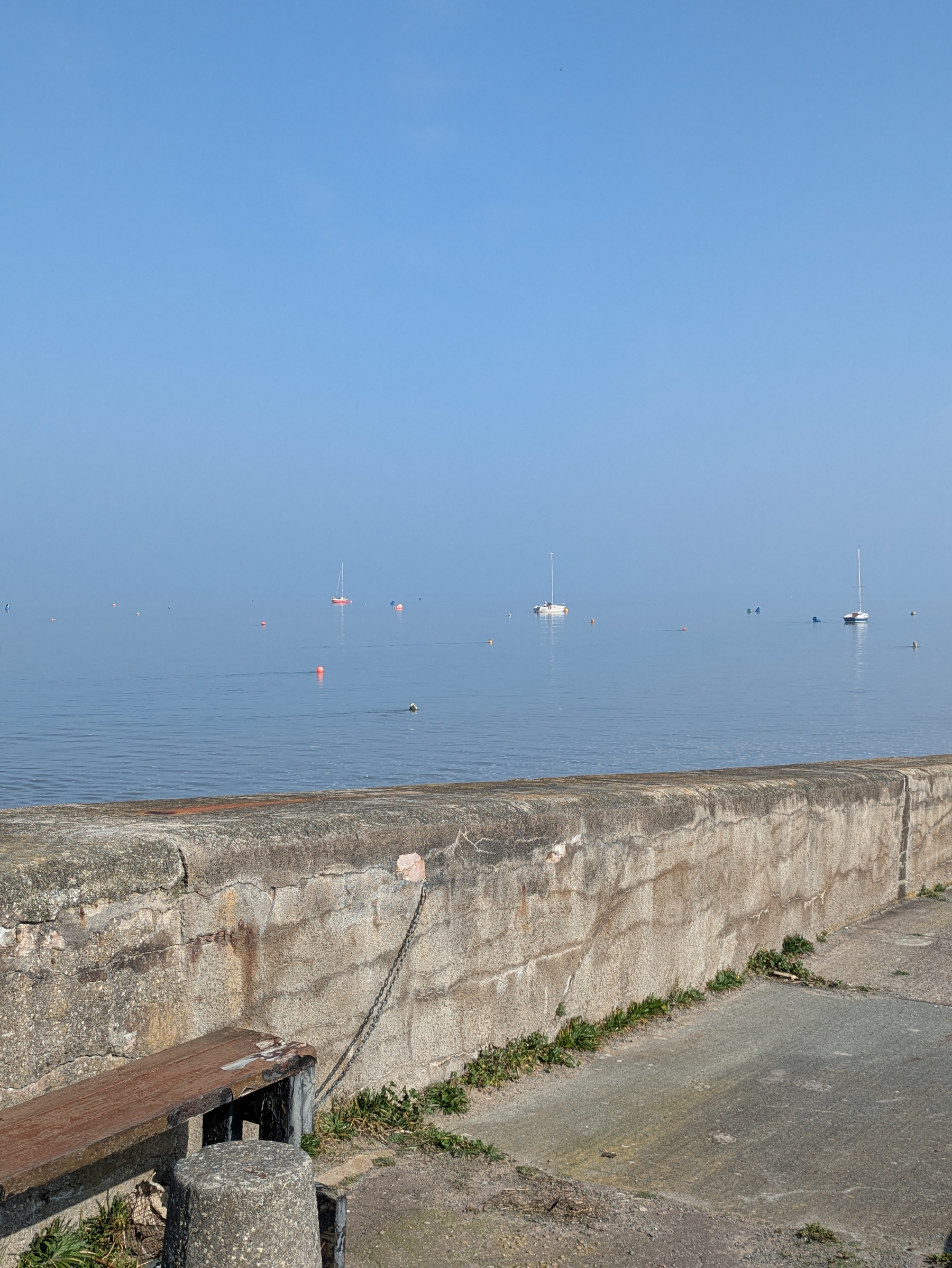 Calm sea with boats floating in the distance under a clear sky, viewed from behind a stone wall.