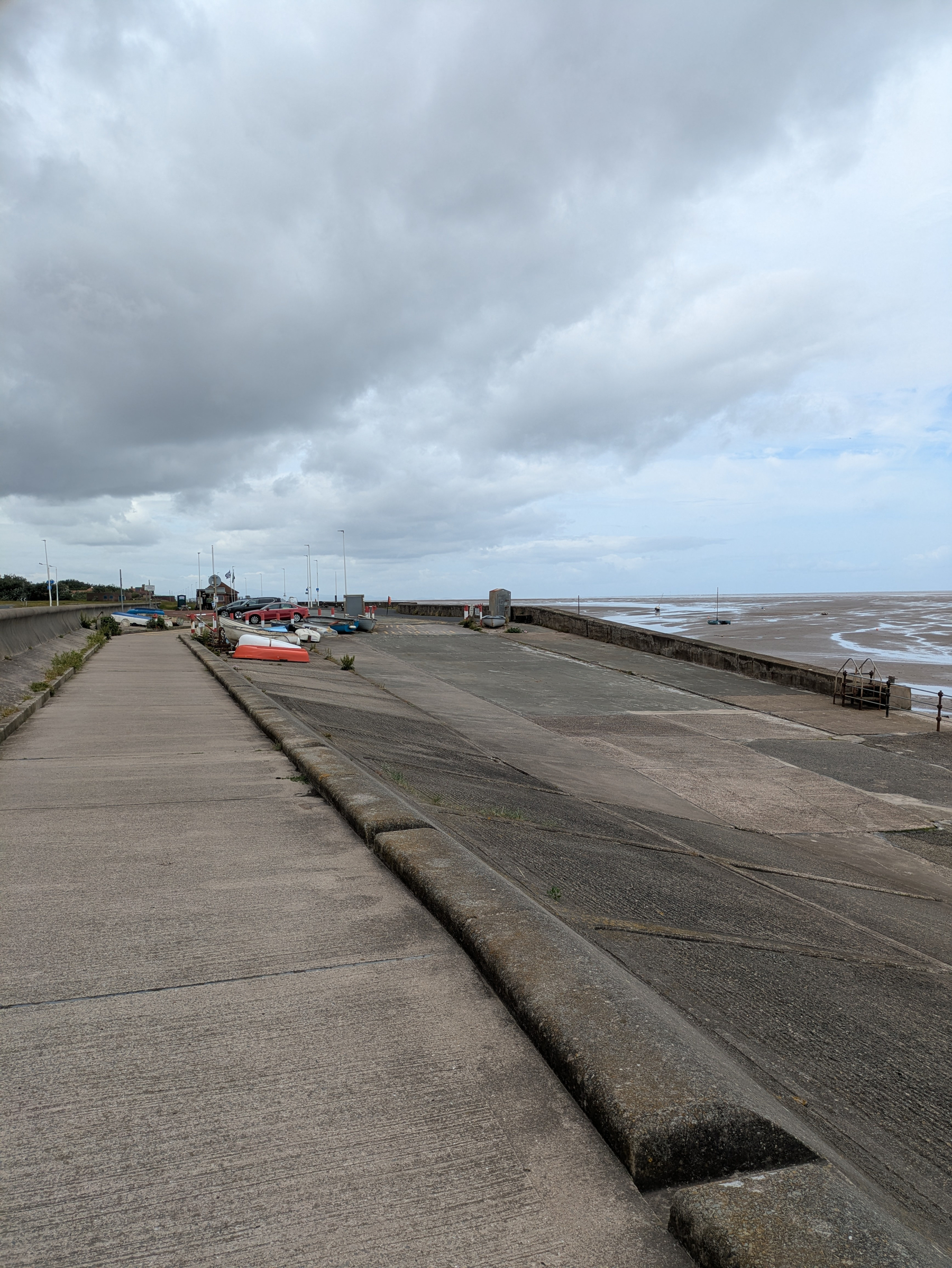 A cloudy coastal scene features a concrete promenade, moored boats, and an expansive sandy beach.
