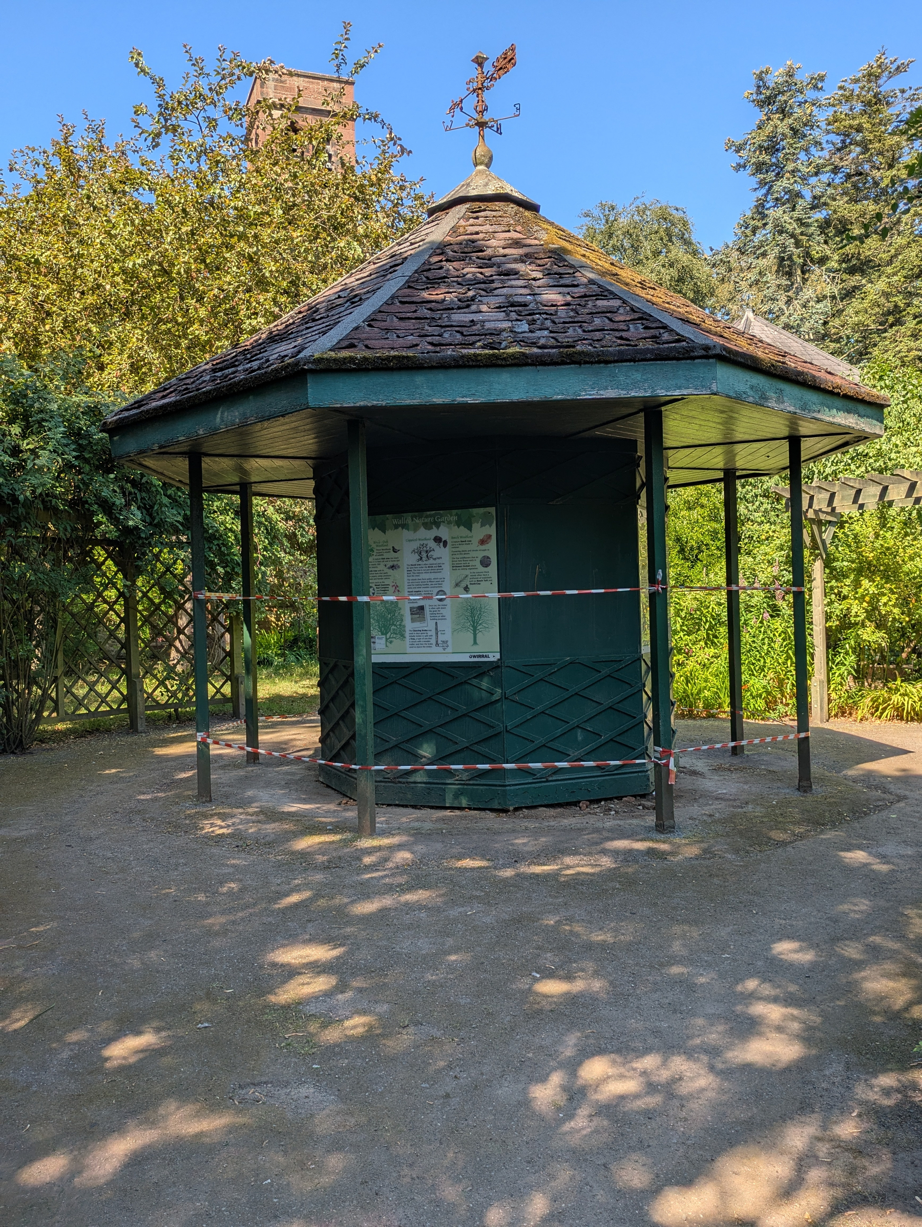 A small wooden gazebo with a weather vane on top is surrounded by caution tape in a garden area.