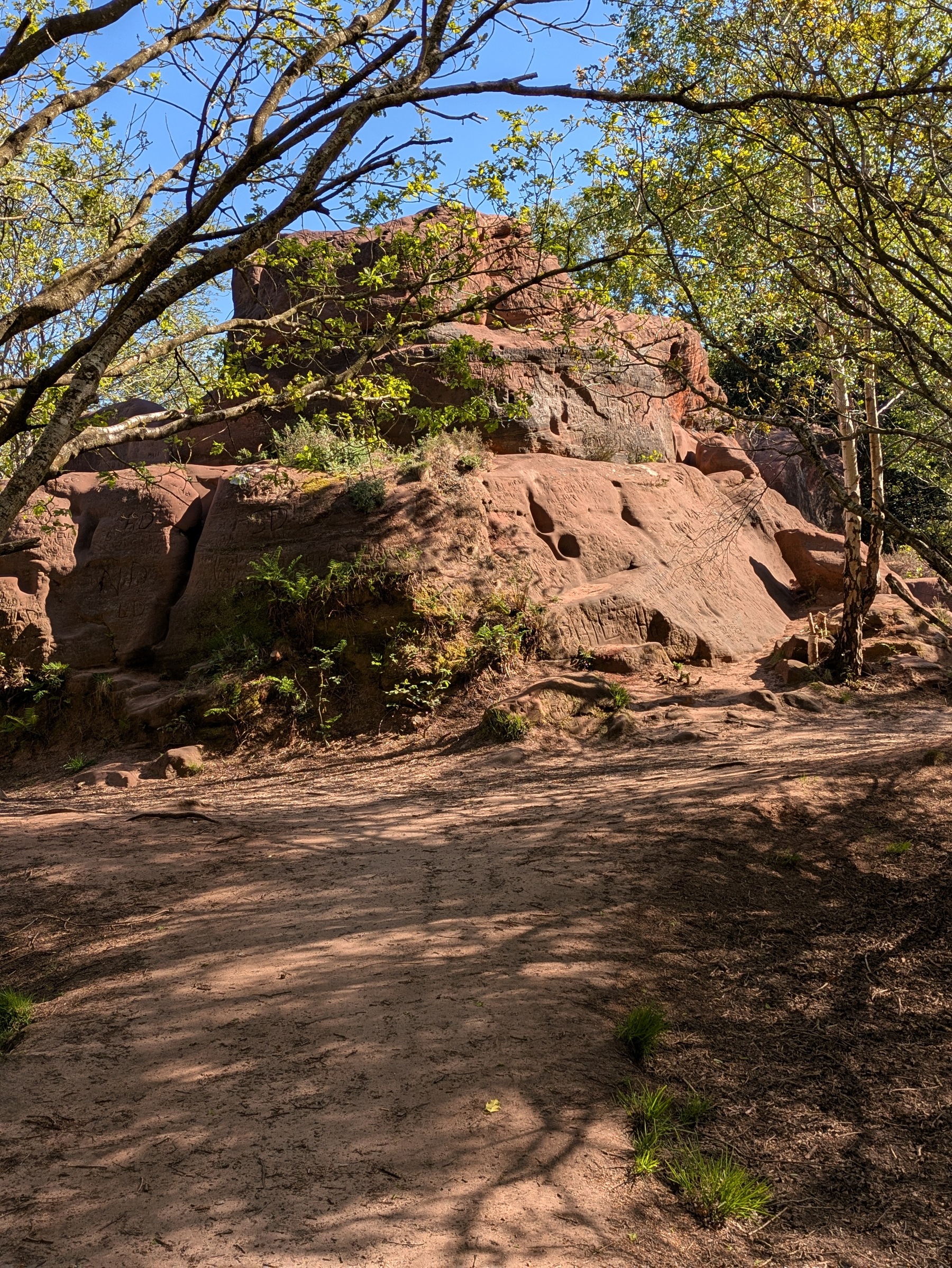 A path leads to a large sandstone rock formation surrounded by trees and foliage.