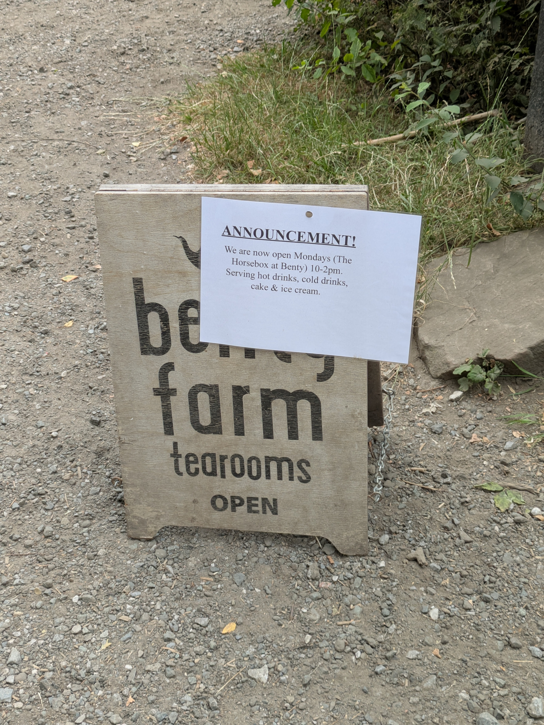 A wooden sign on a gravel path advertises that Benty Farm Tearooms are open and includes an announcement about new Monday hours and offerings.