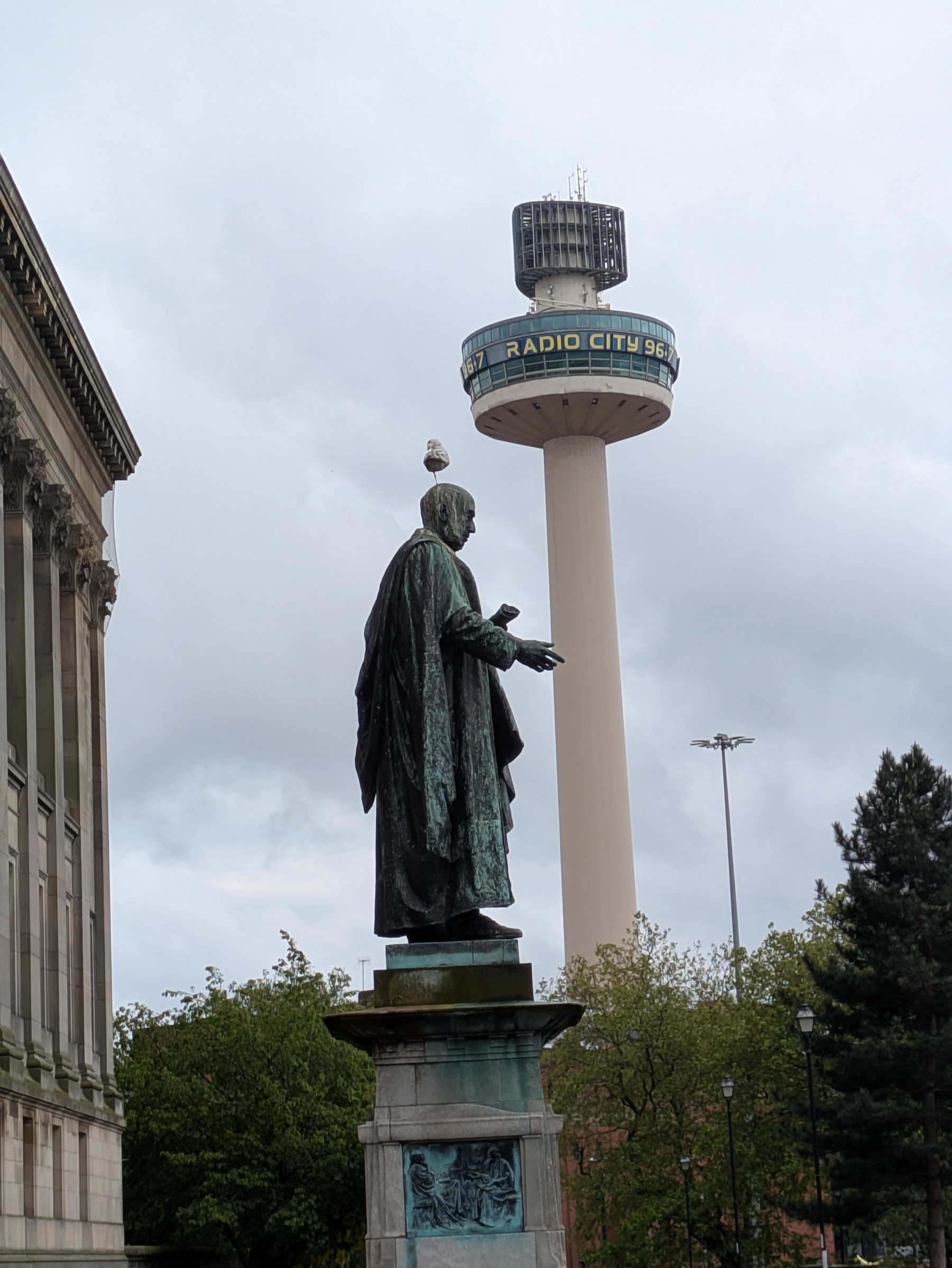 A statue of a historical figure stands near a tall tower with RADIO CITY written on it, beneath a cloudy sky.
