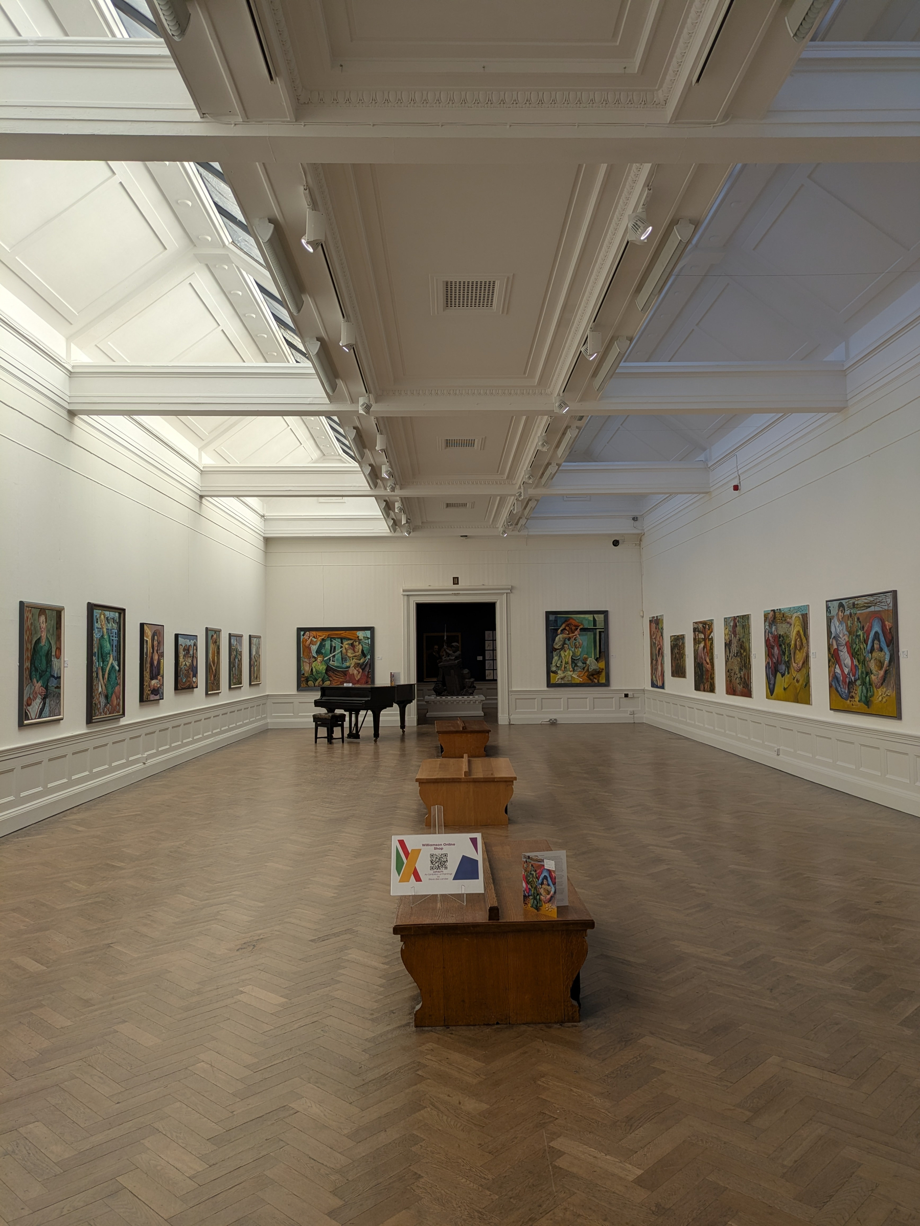 A spacious art gallery with wooden floors features numerous paintings displayed along the walls and benches in the center.