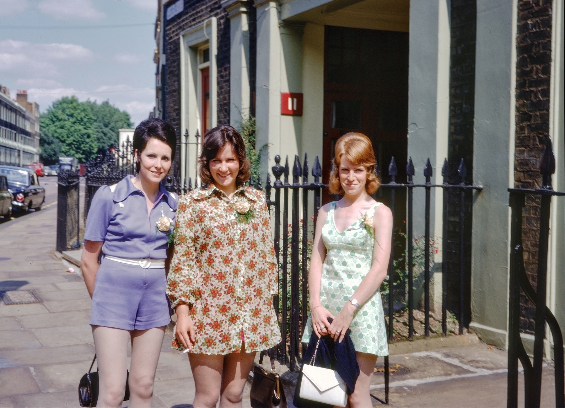 Three women wearing 1960s fashion are standing on a sidewalk in an urban area, each holding a handbag.