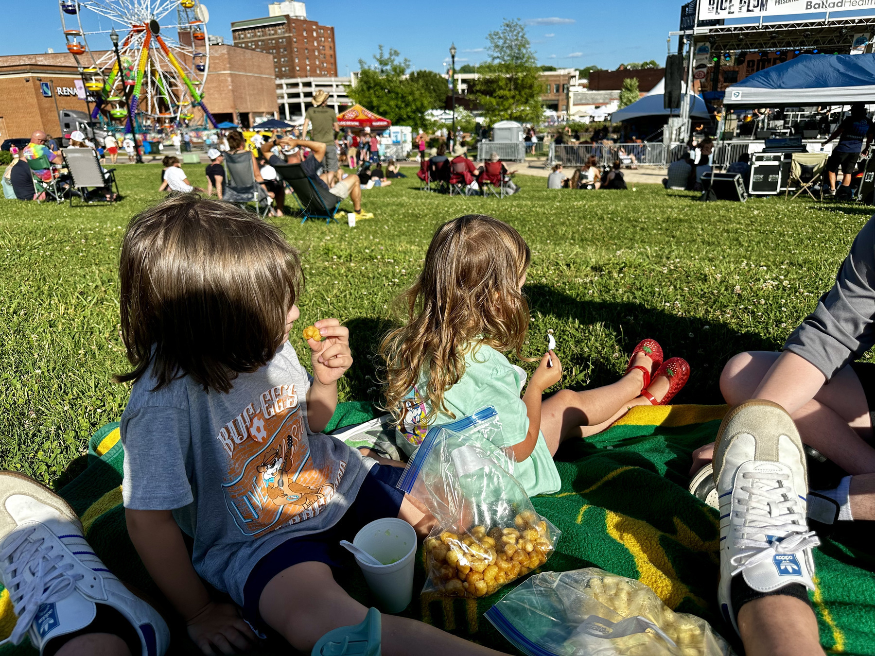 Auto-generated description: Two children are sitting on a blanket eating snacks at an outdoor event with a Ferris wheel in the background.