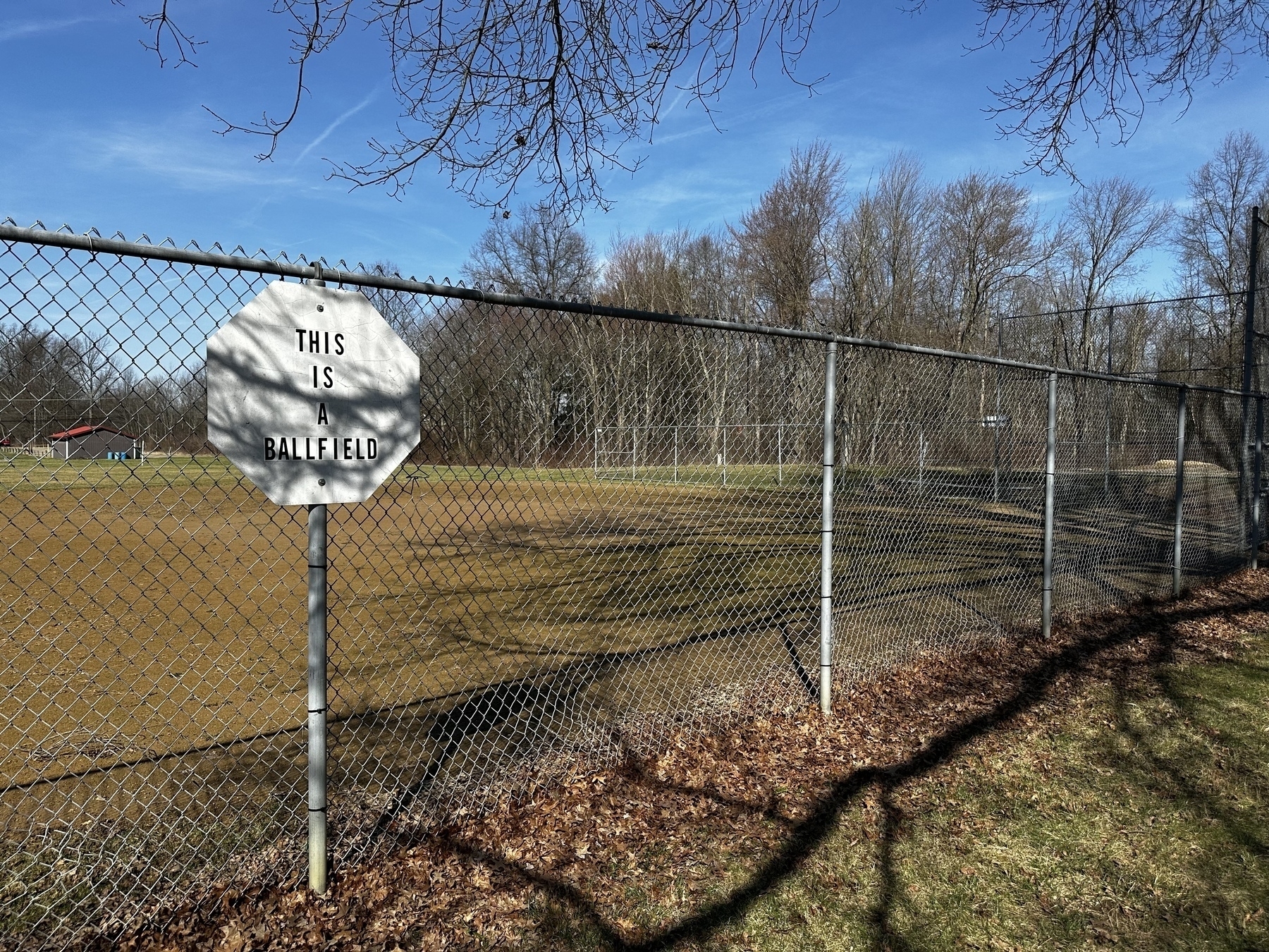 A sign reads "THIS IS A BALLFIELD". It is attached to a chain link fence surrounding a baseball field in a park. 