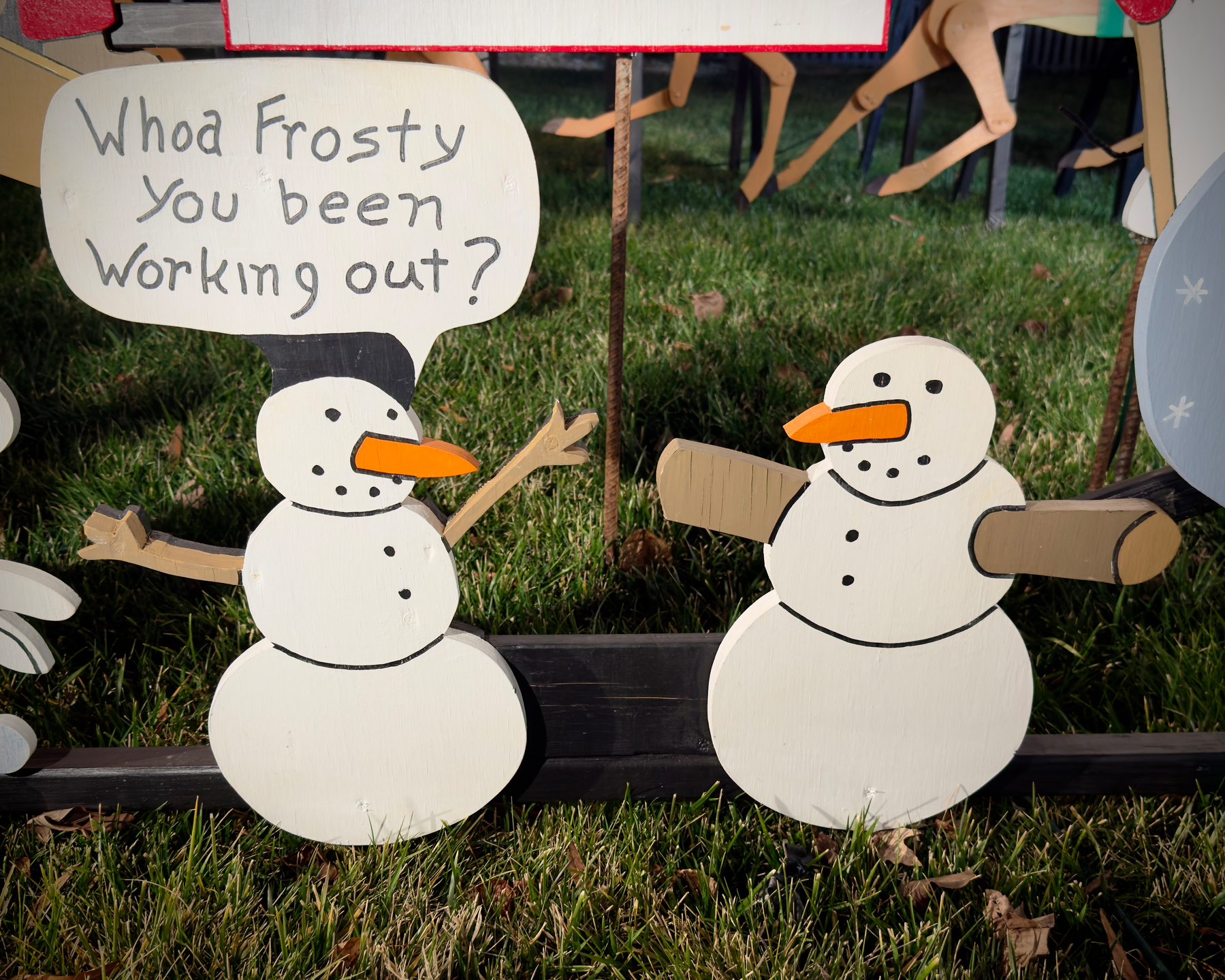 Two wood cutouts of snowmen. One has thin sticks for arms and a word bubble reading, “Whoa Frosty you been working out?” While the other has thick sticks for arms