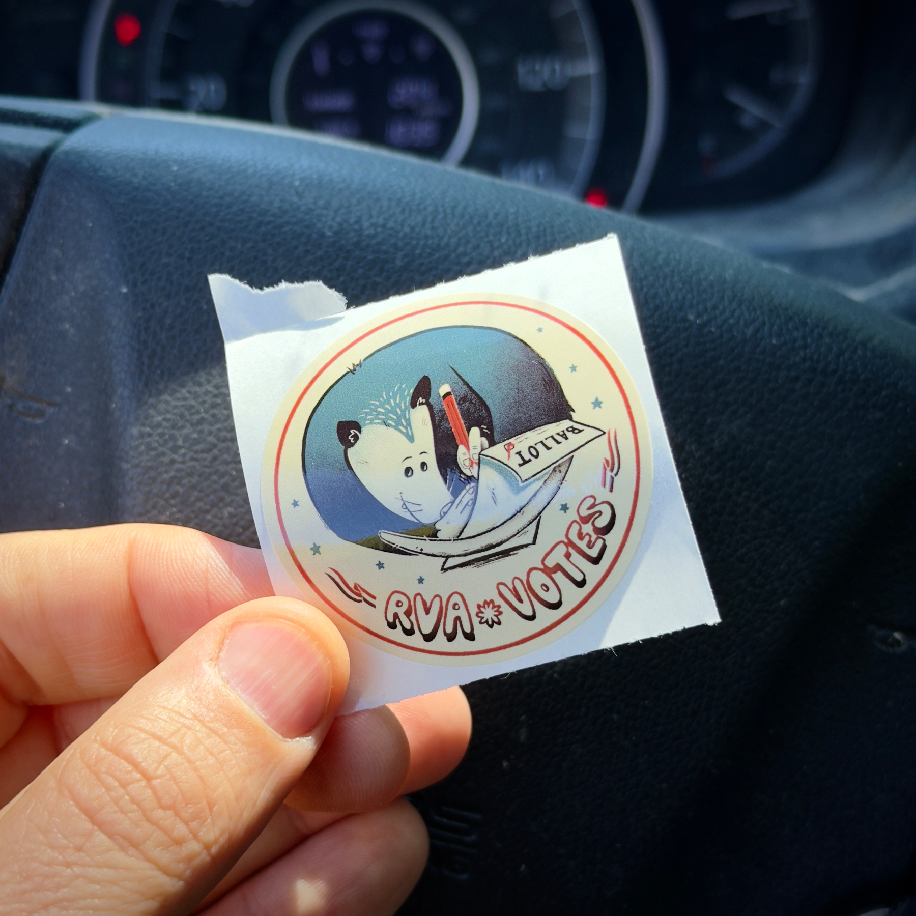 Sticker with a cartoon illustration of a Virginia possum marking on a sheet of paper that says “ballot” with the label “RVA Votes” underneath