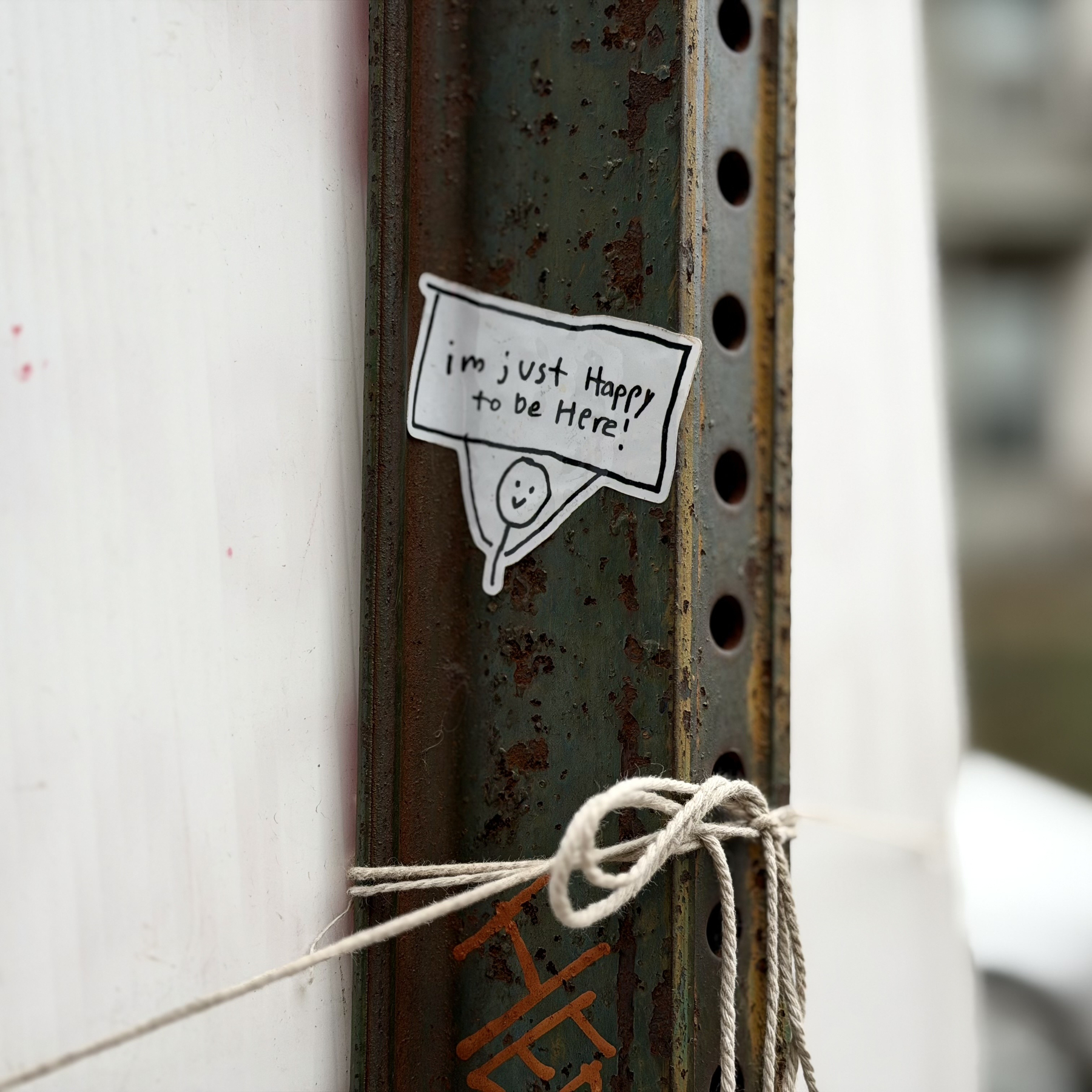 a sticker on a sign post of a stick figure holding a sign that reads "im just Happy to be Here!"
