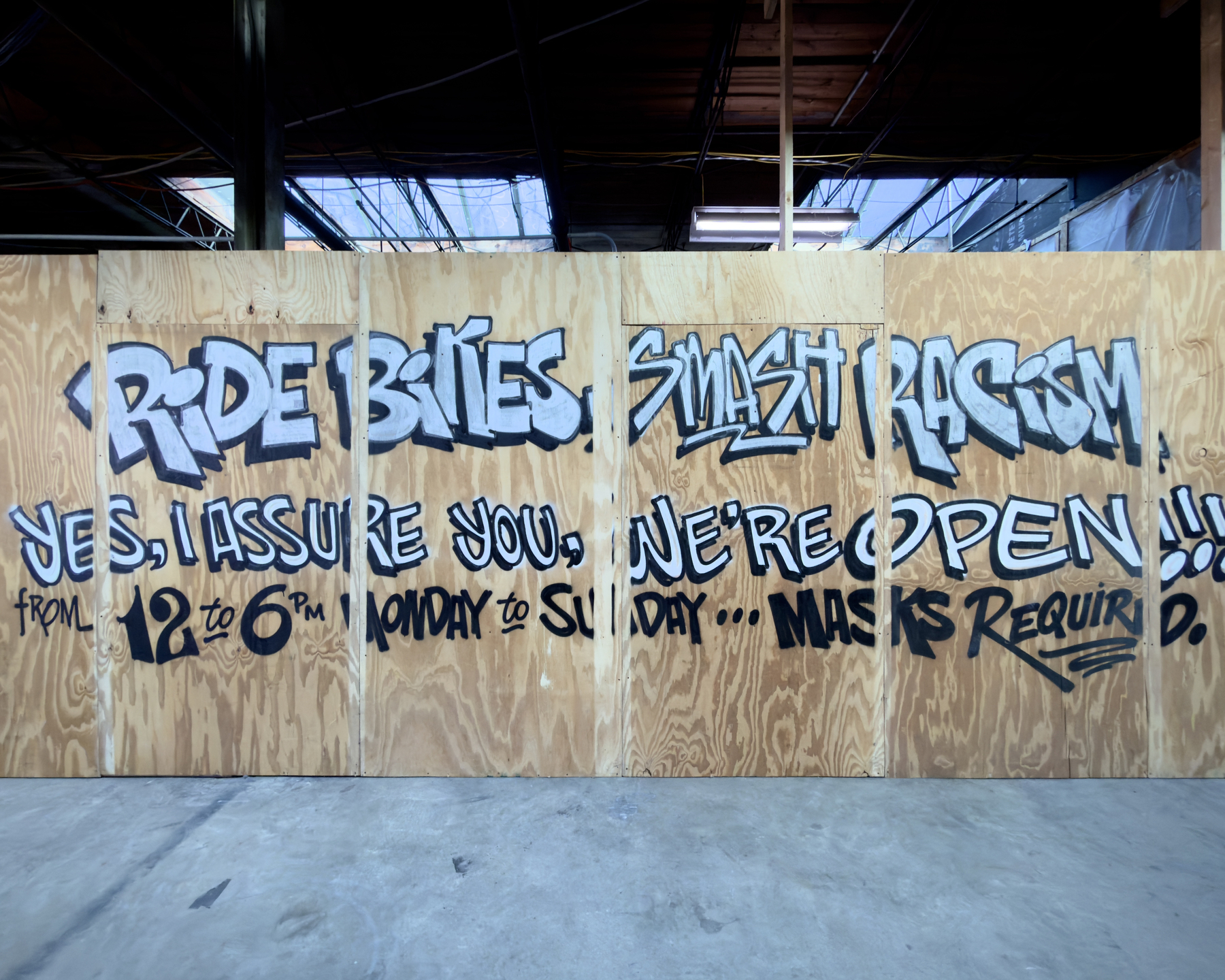 graffiti style painted words on plywood wall reading 'ride bikes smash racism' on the first linen and 'yes I assure you we're open' on the second line