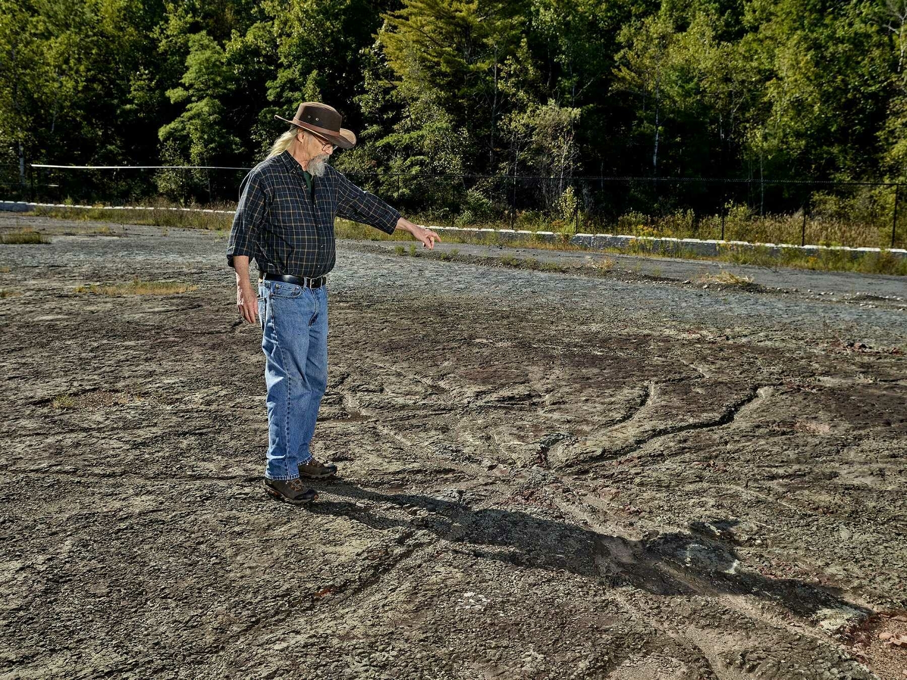 A photograph of a white-haired man in a hat gesturing at the fosilised remains of a tree root system on baren rocky ground. Photo by Noah Kalina.