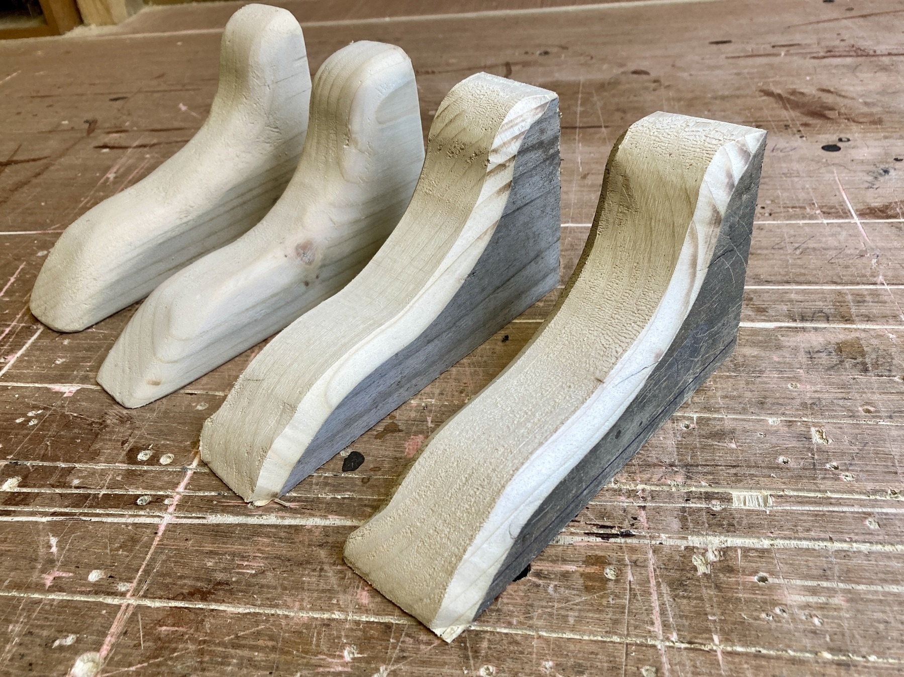 Four wooden shelf brackets on a workbench. The two inthe foreground are roughly shaped before sanding.