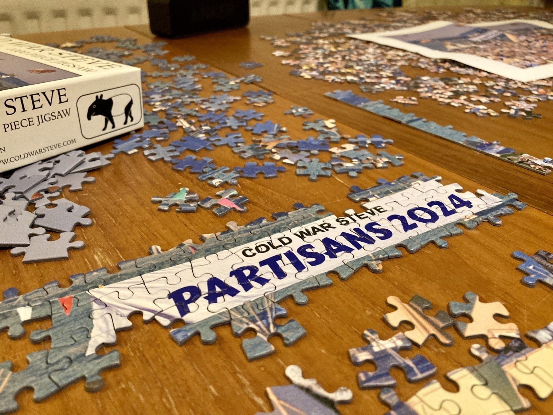 The beginning of the process of doing a jigsaw. One small section is complete, a banner reading Cold War Steve Partisans 2024. Various pieces, a box and a sheet of paper are visible on the table. 