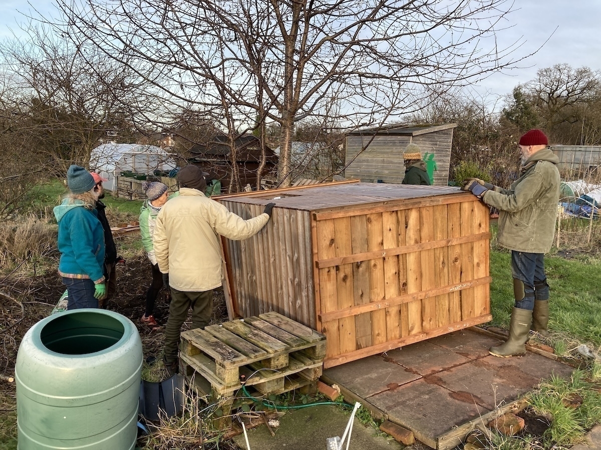 A garden shed which has fallen backwards so the base is facing the camera. A group of people in winter coats and hats is gathered around it, ready to lift. 