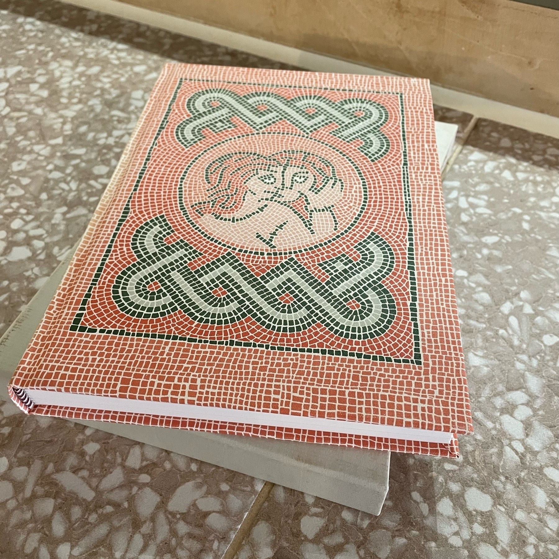 Hardback book with cover in the form of a full red and green mosaic showing two figurative lovers. Book sits atop a plain cream coloured cardboard sleeve. 