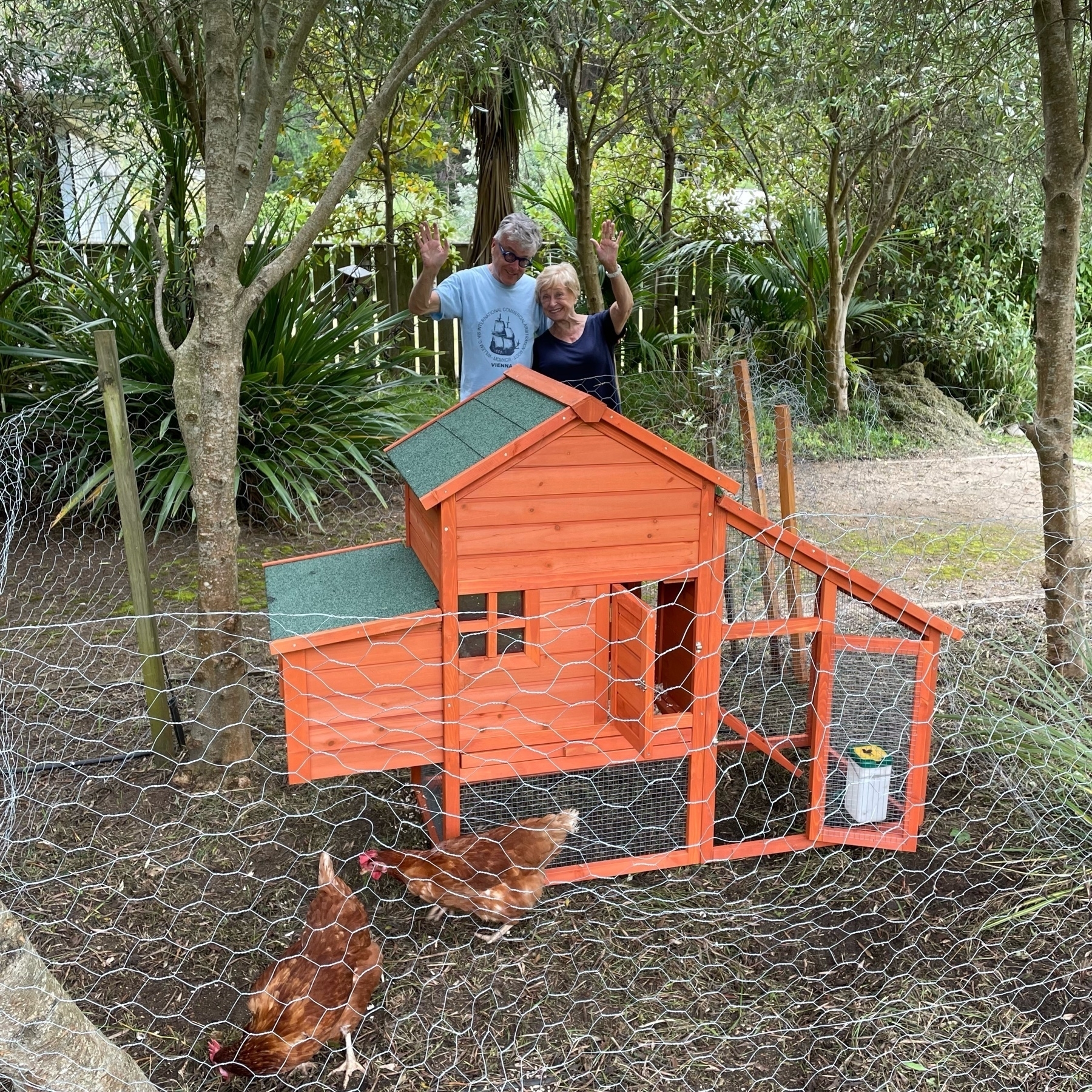 Chicken coop with two people waving. Two chickens in the foreground.
