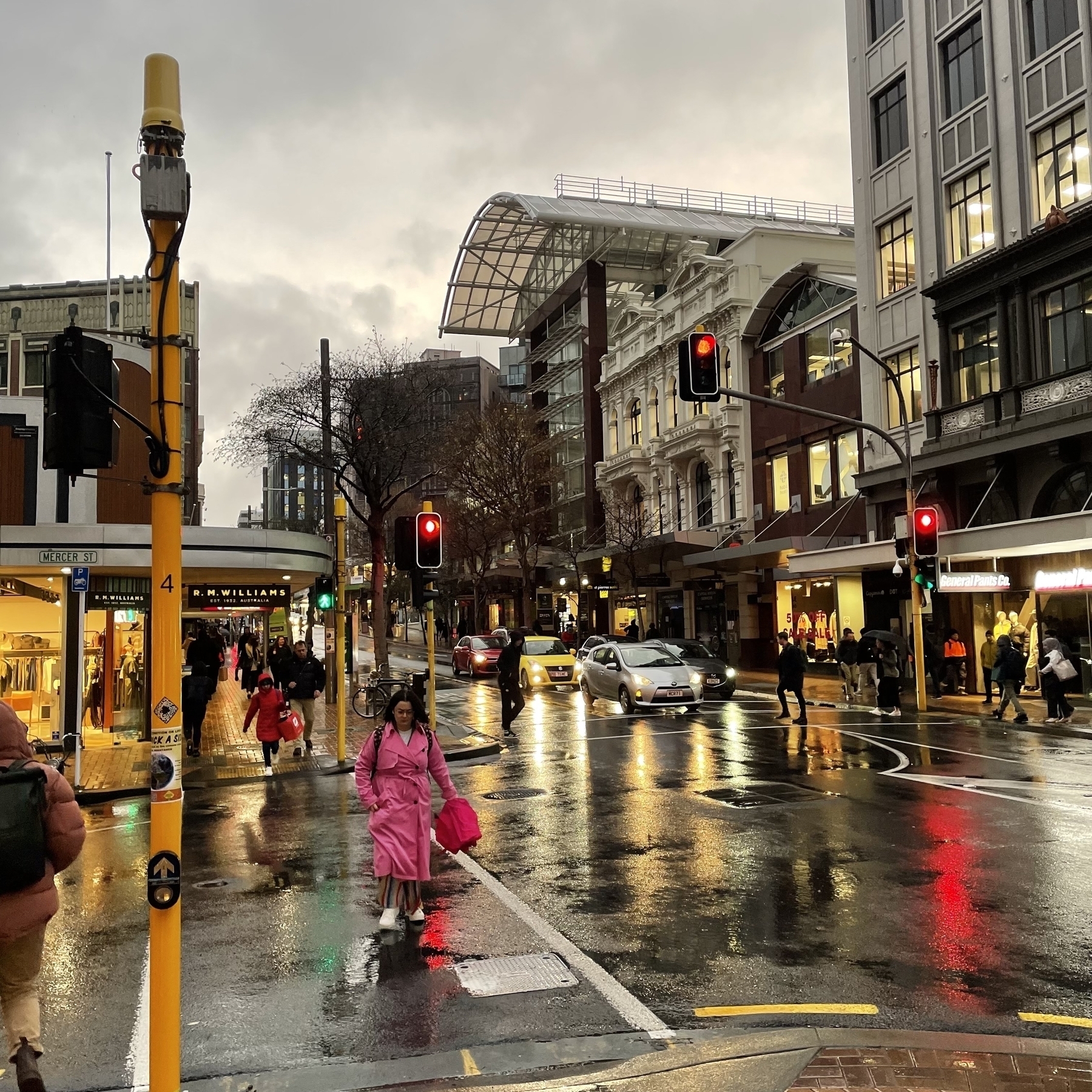 Rain washed street scape with yellow and red lights from shops, cars and street lights reflecting on the street. People in pink, red and other colours cross the road. 