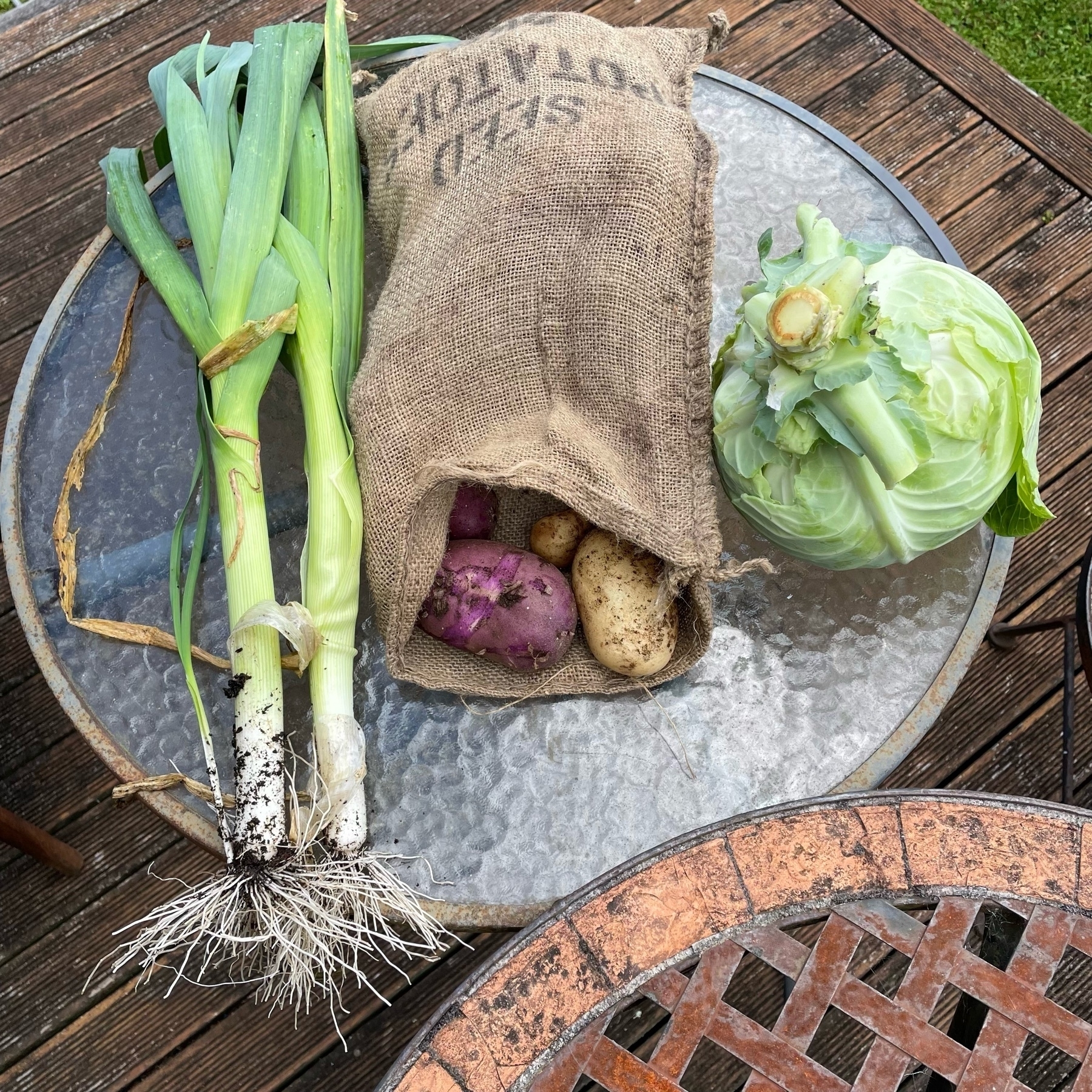 Leeks, multi-coloured potatoes spilling out of a hessian bag, and a cabbage on a rustic table. 