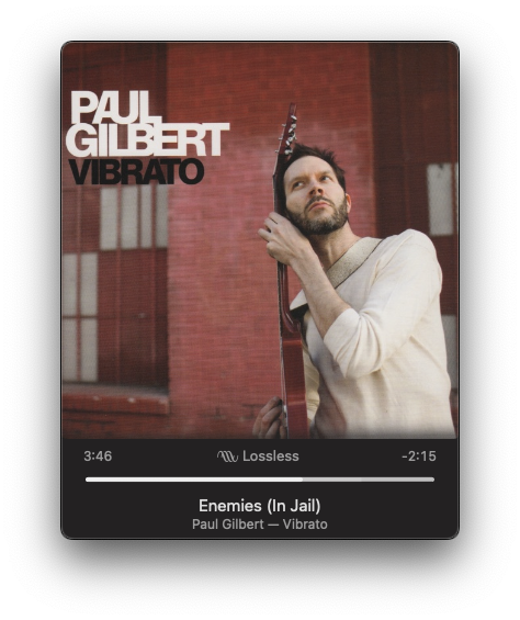 A man is holding a guitar next to a red brick wall with the text Paul Gilbert Vibrato on the upper left.