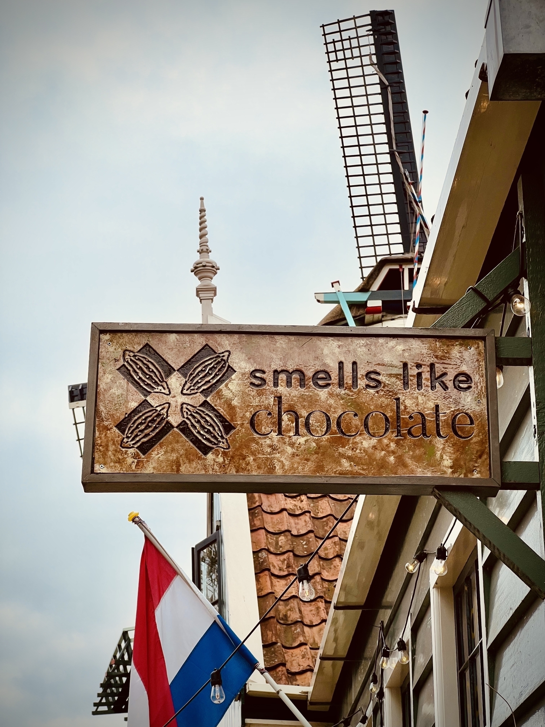 Sign saying “smells like chocolate”, with part of a windmill and the Dutch flag in the background 