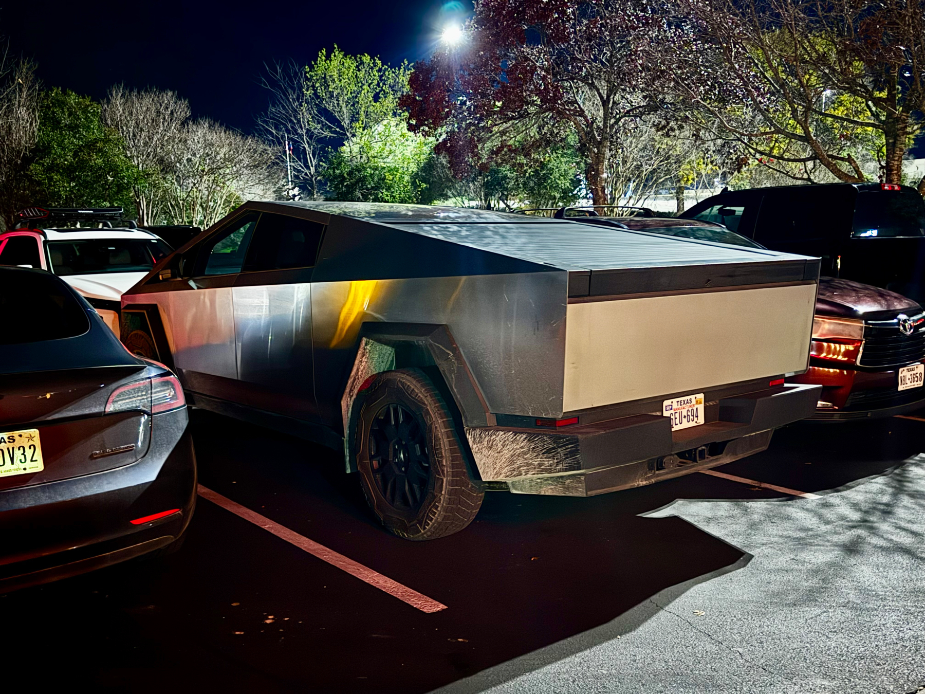 A Tesla Cybertruck parked in a parking lot at night, surrounded by other vehicles. The Cybertruck&rsquo;s distinct angular design and metallic finish are visible. The photo is taken from a three-quarter rear perspective.