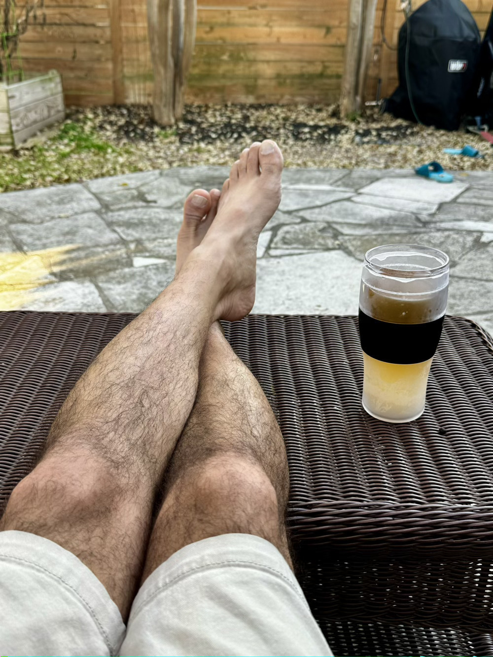 Person&rsquo;s legs stretched out on a wicker surface, with a glass of beer nearby, in a garden setting.