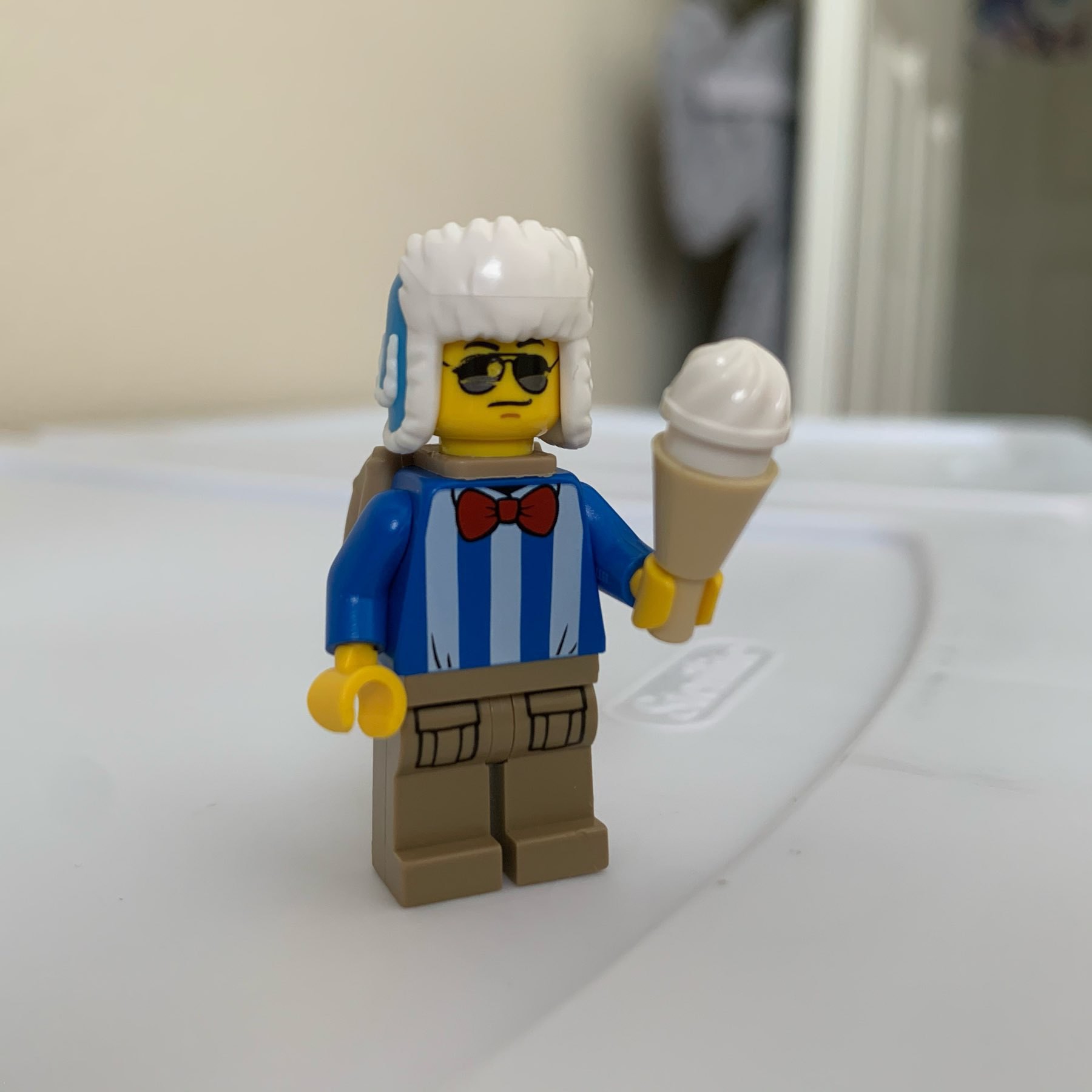 LEGO minifigure wearing blue shirt, red bowtie, sunglasses, backpack, winter hat, and holding an ice cream cone.