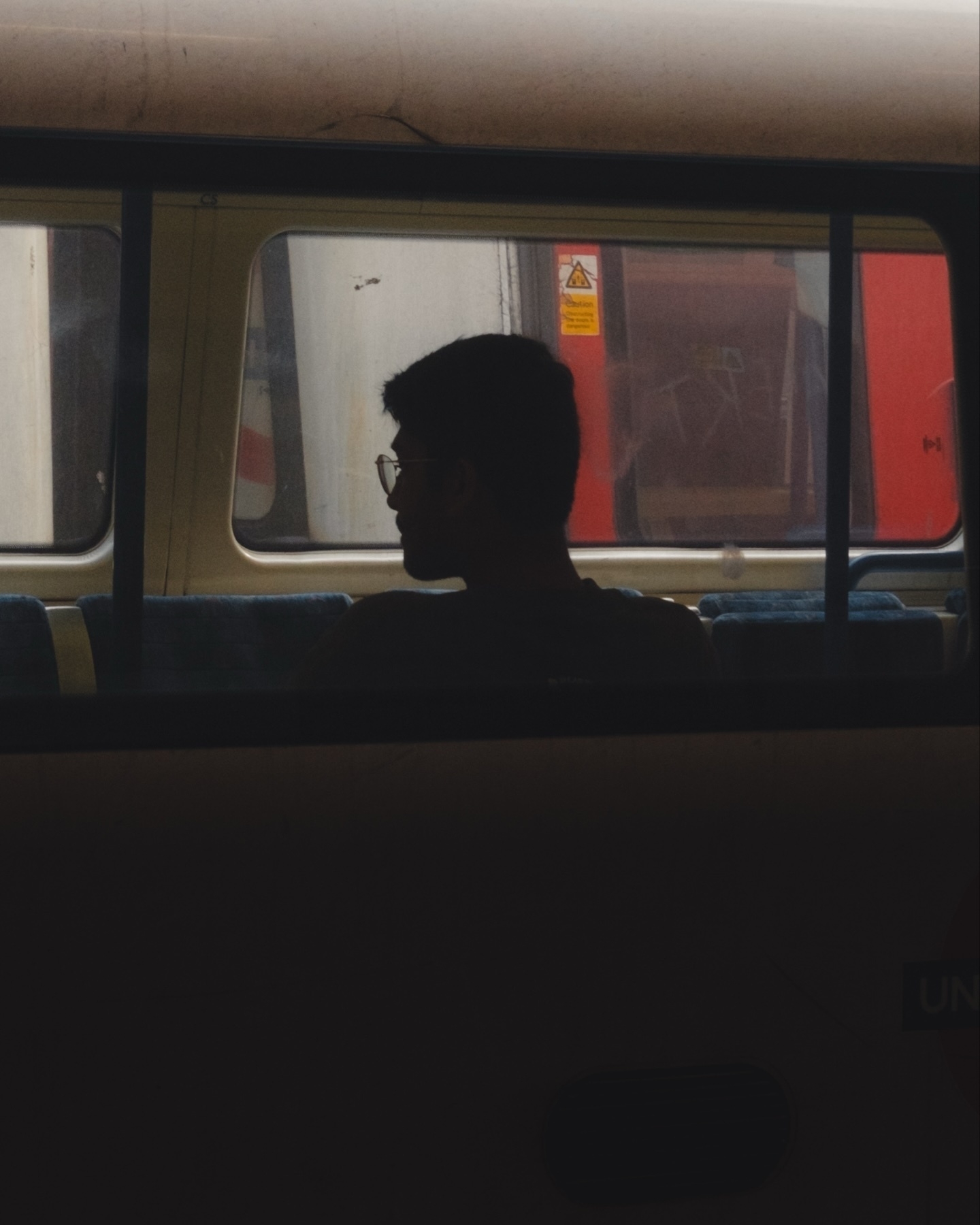 A silhouette of a person with glasses seated inside a train, looking out of the window. The background shows another train outside with some graffiti and a warning sign. The overall scene has a dimly lit, contemplative mood.