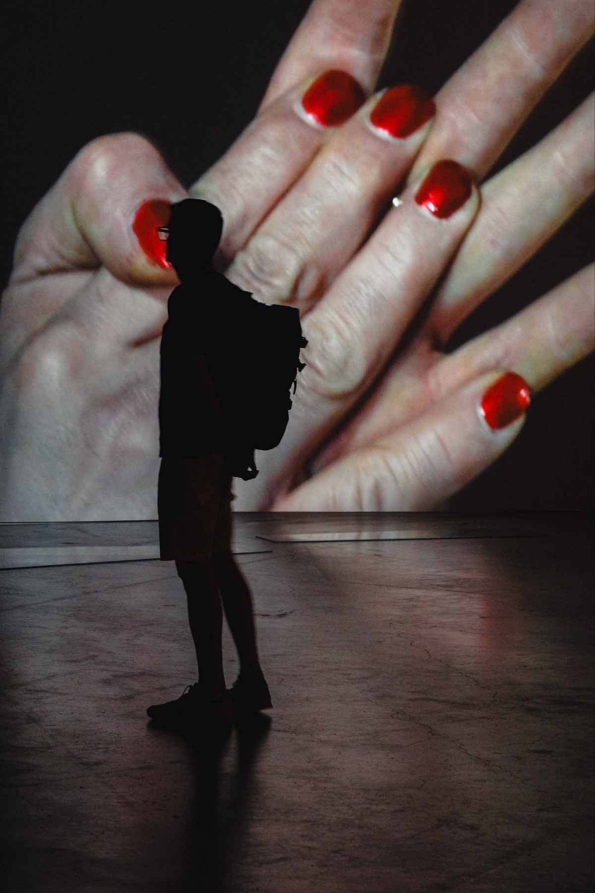 A silhouette of a person with a backpack stands in front of a large projected image of a hand with red nail polish. The setting appears to be an indoor space with a concrete floor.