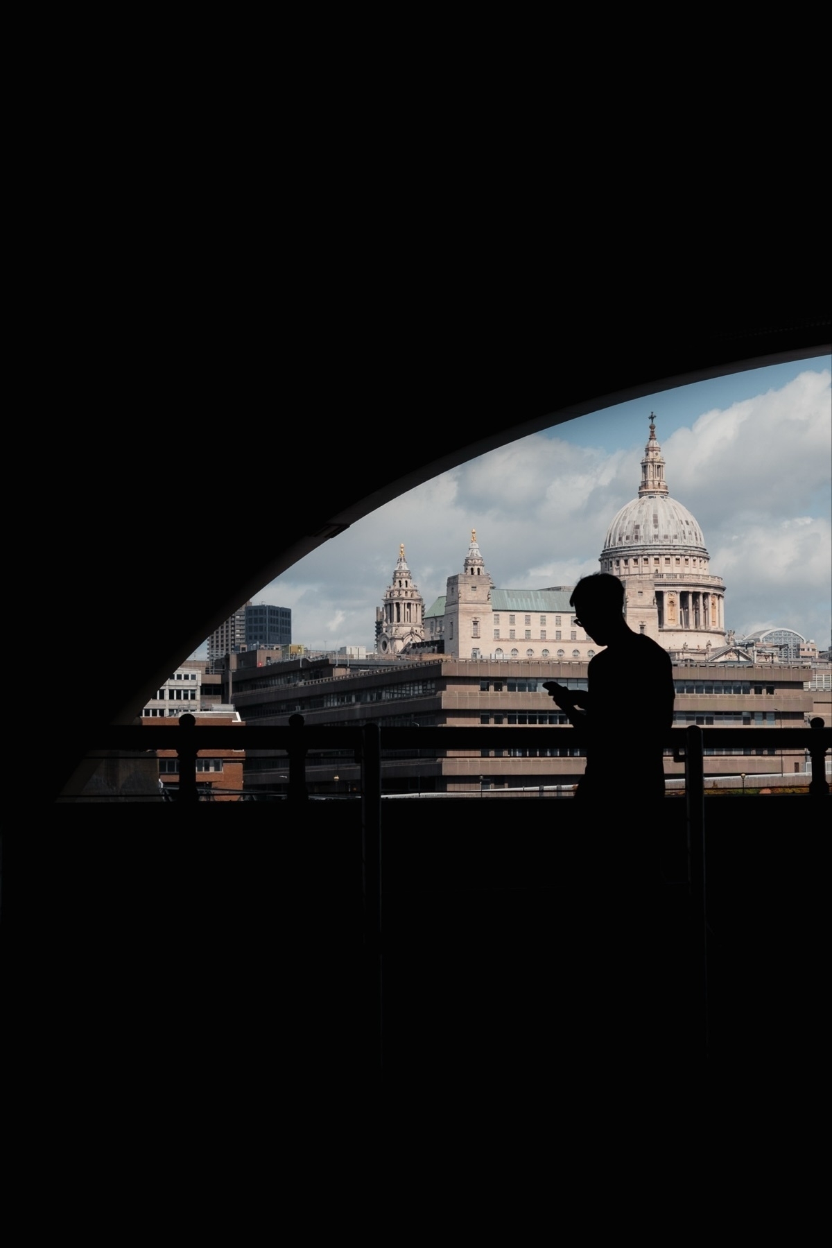 A silhouette of a person looking at a smartphone is framed against the backdrop of historic buildings, including a prominent domed structure under an archway. The scene features a mix of classical and modern architecture under a partly cloudy sky.